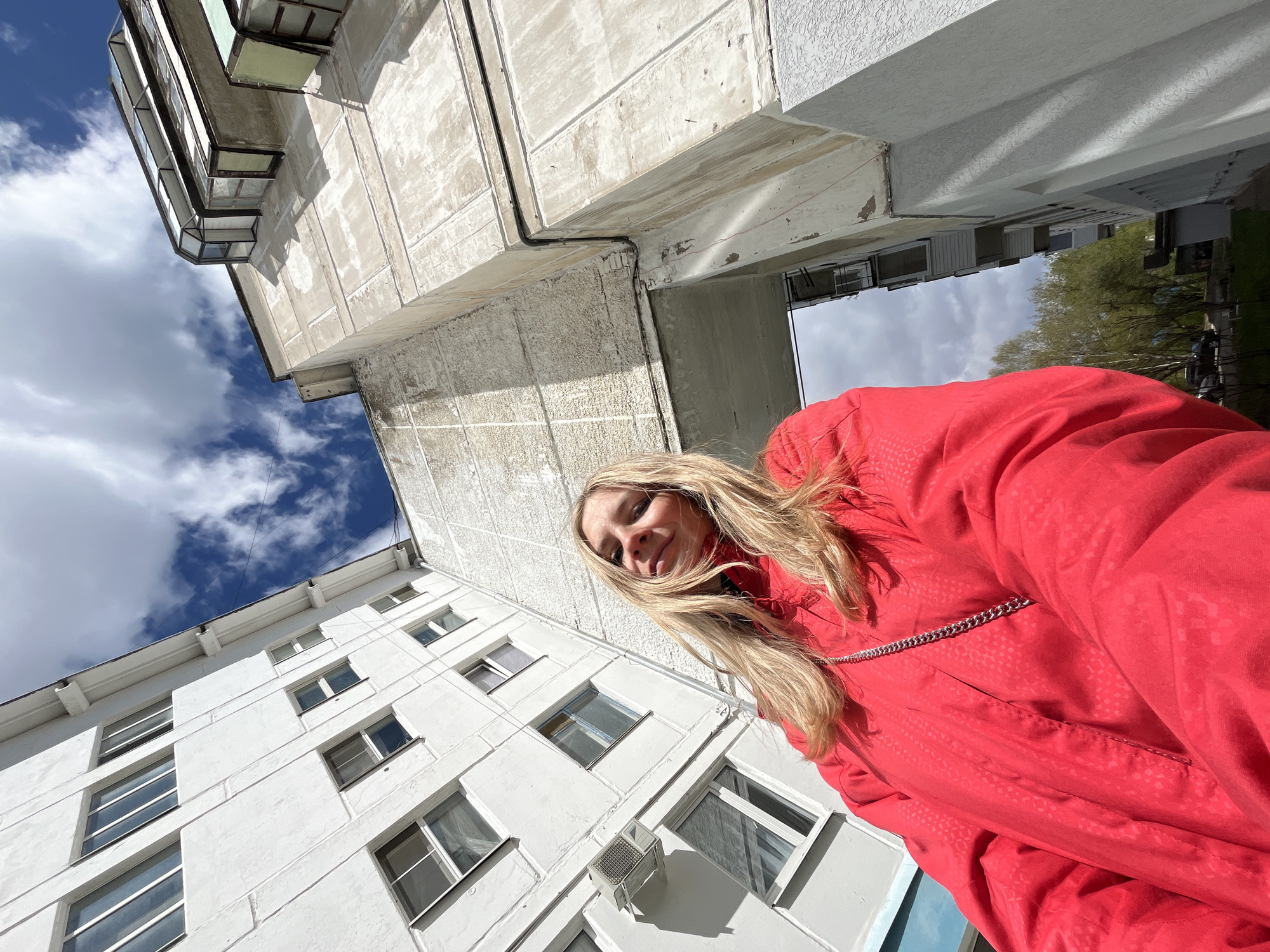 A young woman with blonde hair is standing in front of two high-rise buildings under a partly cloudy sky. She is wearing a red jacket. The buildings appear to be residential apartments, with multiple windows visible on their facades. One building is white, and the other is grey. The photo is taken from a low angle. The location is in the city of Chaikovsky, Russia.