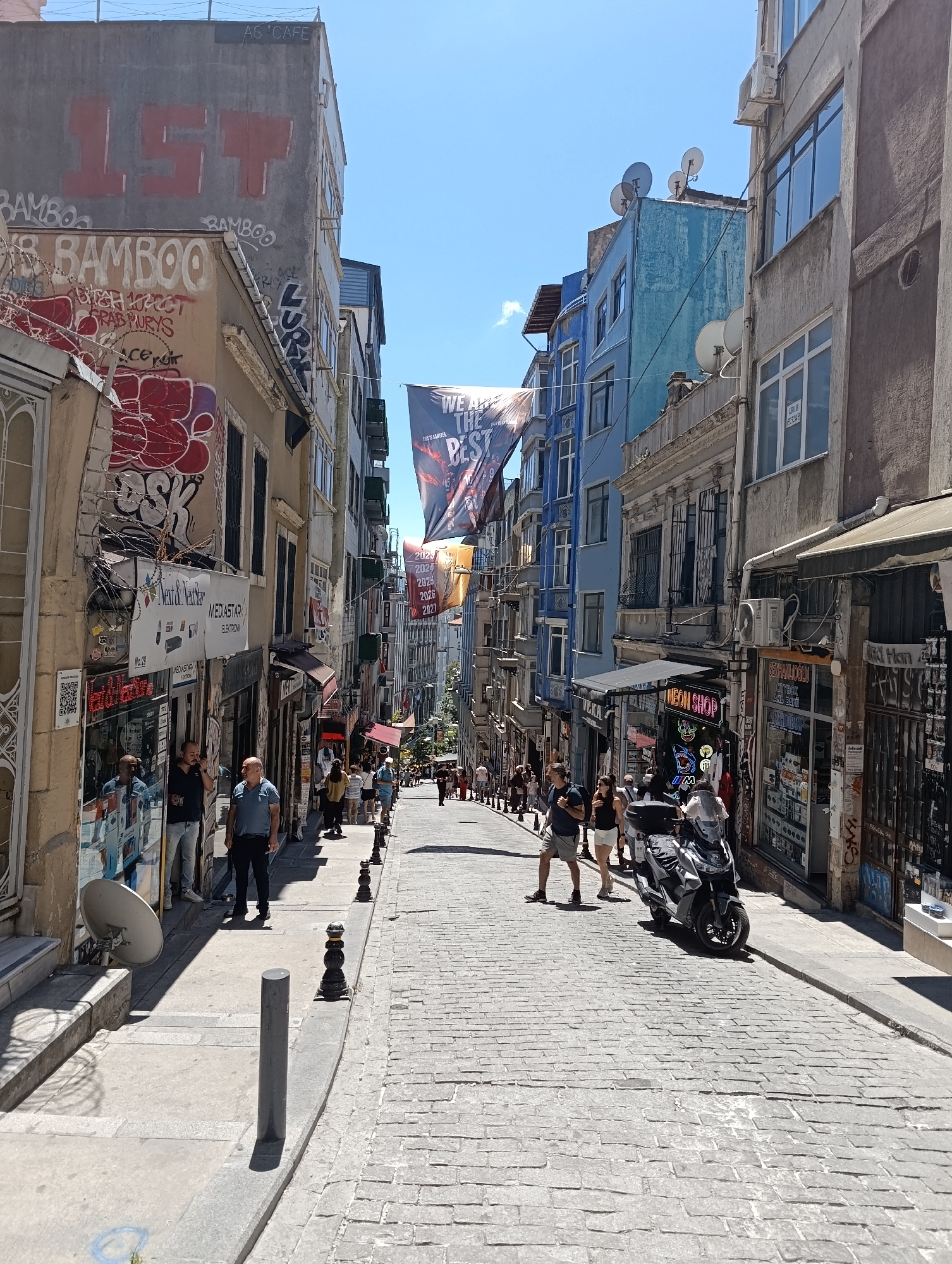 This is a wide, eye-level, outdoor shot of a narrow, uphill cobblestone street in Beyoğlu, Turkey, lined with multi-story buildings. The sky is clear and blue. The buildings on either side of the street have an eclectic mix of architectural styles and are adorned with numerous satellite dishes. The left side of the street features buildings with graffiti, including large red "1ST" and "BAMBOO" lettering. Shops with open storefronts are visible, displaying goods. People are walking up the street, some in groups and some individually. On the right side of the street, a dark-colored scooter is parked. The street is illuminated by bright sunlight, casting shadows on the cobblestones and pavement.