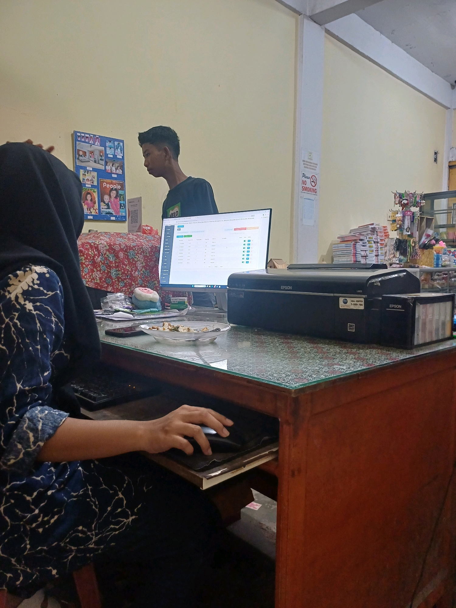 In a room in Dumai City, Indonesia, a young male with dark hair is standing and looking at a computer screen. A person wearing a dark hijab and a blue and white patterned garment is seated at a desk in front of the computer, using a mouse. The desk is made of wood and has a glass top. On the desk, there is a computer monitor displaying a spreadsheet, a black Epson printer, a plate with food, and various papers. A "Please No Smoking" sign is visible on the wall. The room appears to be an office or a shop, with shelves containing products in the background.