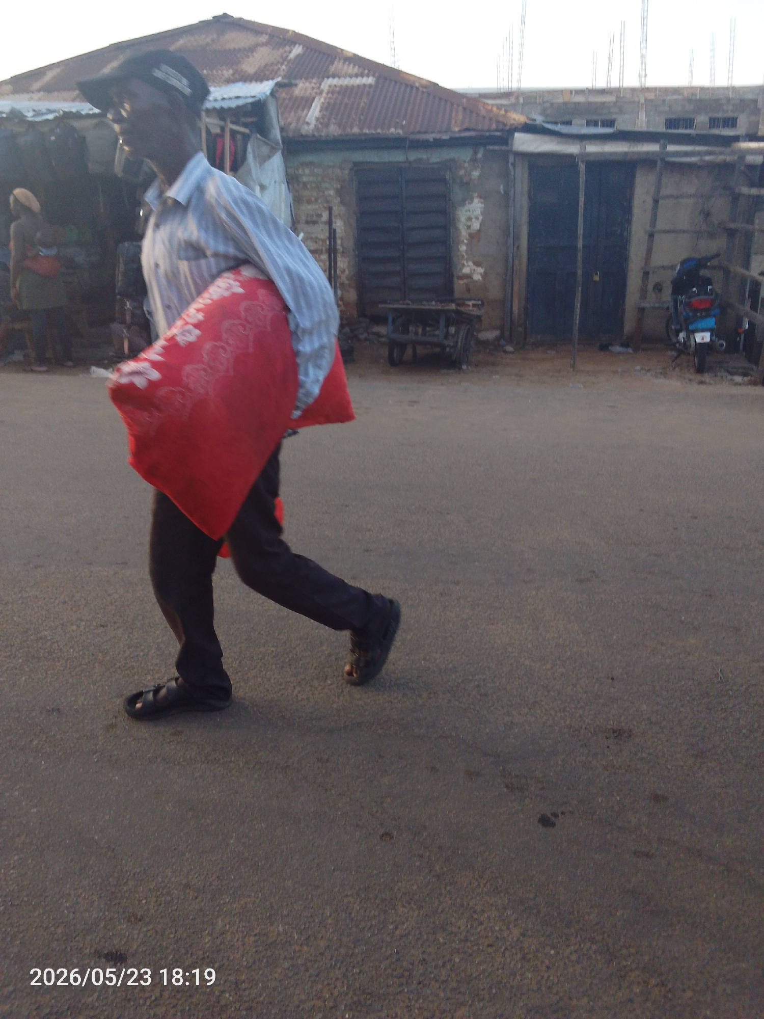 In Lokoja, Nigeria, a man walks along a paved road carrying a large, red bag with a floral pattern over his shoulder. He is wearing a striped shirt, dark pants, a cap, and sandals. The background includes dilapidated buildings with corrugated iron roofs, one of which has dark doors and barred windows. A motorcycle is parked near a building with a ladder leaning against it. Other figures are visible near what appears to be a market stall in the background. The time stamp on the image indicates 2026/05/23 18:19.