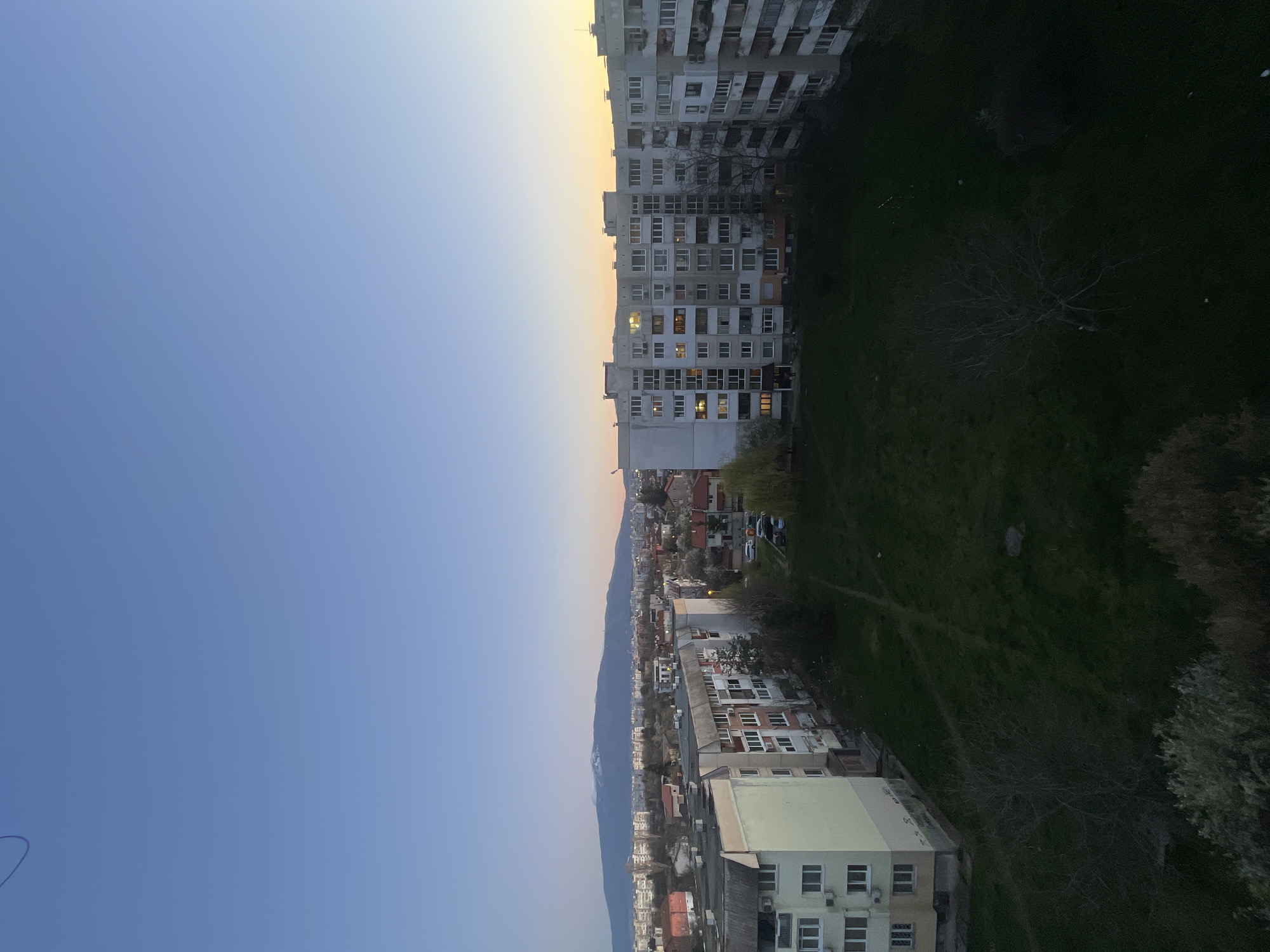 This image depicts an urban scene, likely from a high vantage point, possibly in Sofia, Bulgaria. A large apartment building stands on the right, adjacent to a grassy area with a tree. Below, a residential neighborhood extends toward the base of a mountain visible in the distance. The sky transitions from blue to a yellowish hue, suggesting either dawn or dusk. There is no presence of people, actions, or direct interactions.