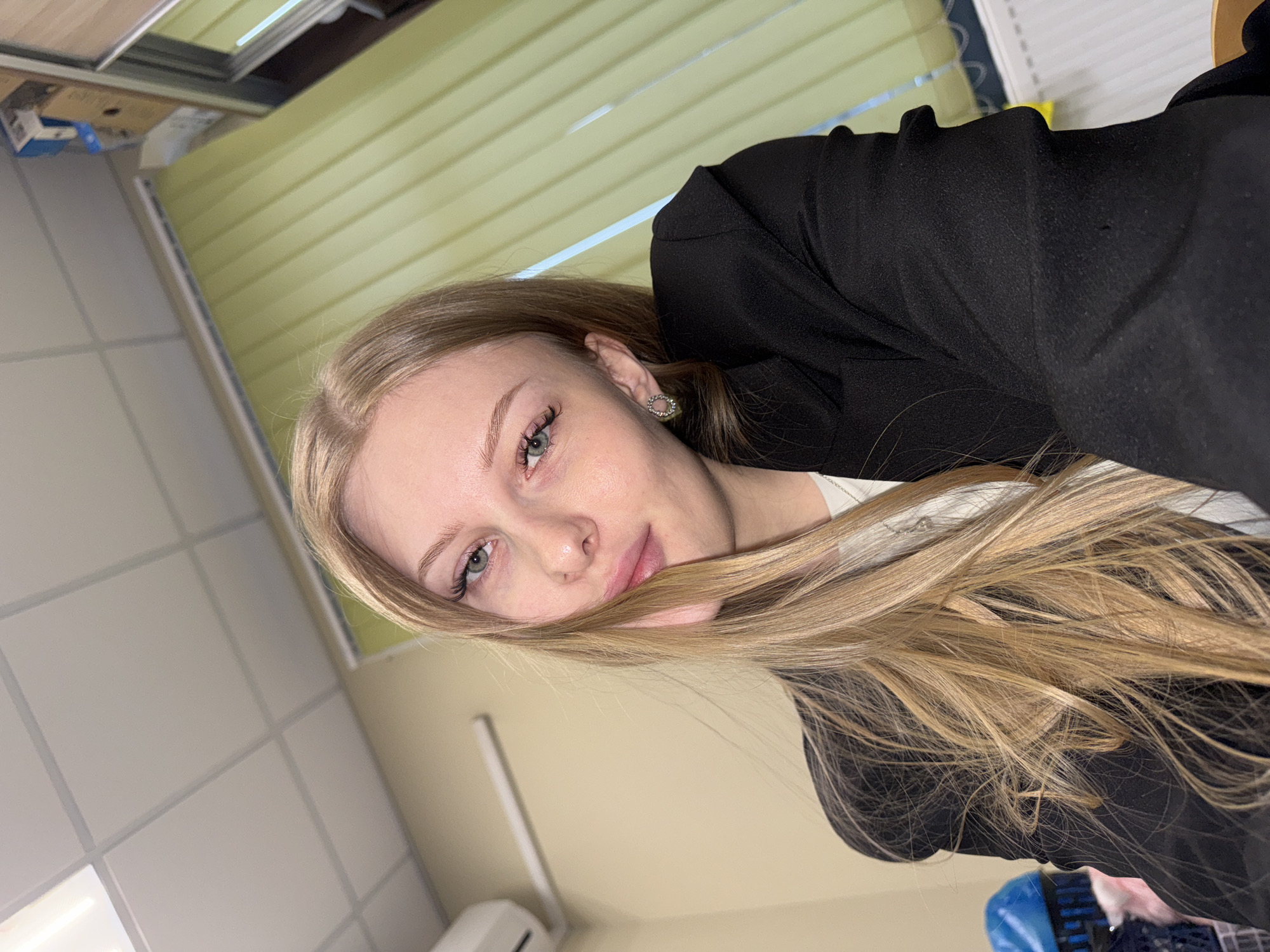 A fair-skinned young woman with long blonde hair and green eyes is looking at the camera. She is wearing a black blazer over a white shirt. Her hair partially obscures her face. An earring is visible in her left ear. The background consists of a horizontally-striped, light-green window blind and a drop ceiling with rectangular tiles. A white air conditioning unit is visible in the lower left corner. Based on prior metadata associated with similar images, the location of this photograph is likely Perm, Russia.
