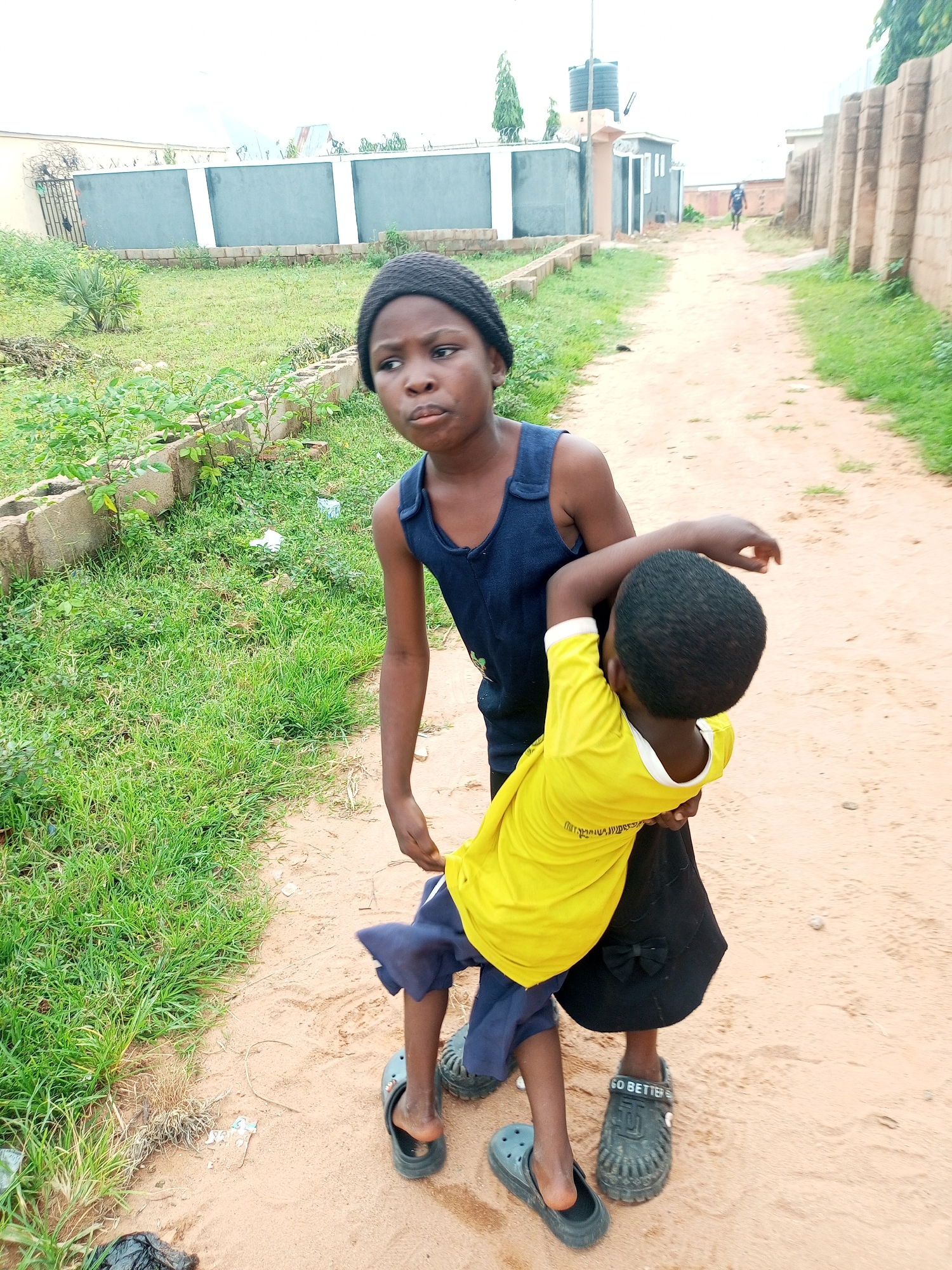 Two young children are standing on a dirt path in what appears to be a residential area. The child on the left, facing the camera, wears a dark blue sleeveless top and a black knitted beanie. This child is looking slightly upward and to the left with a serious expression. The child on the right, facing away from the camera, wears a bright yellow t-shirt and a black skirt with a bow detail. This child is embracing the child on the left from behind. Both children are wearing dark-colored slip-on shoes. The path is bordered by dry grass and some low-lying plants on the left, and a brick wall on the right. In the background, there are buildings, including a long wall with light-colored sections and a fence, and further down the path, another building with a water tank on top. The sky is overcast.