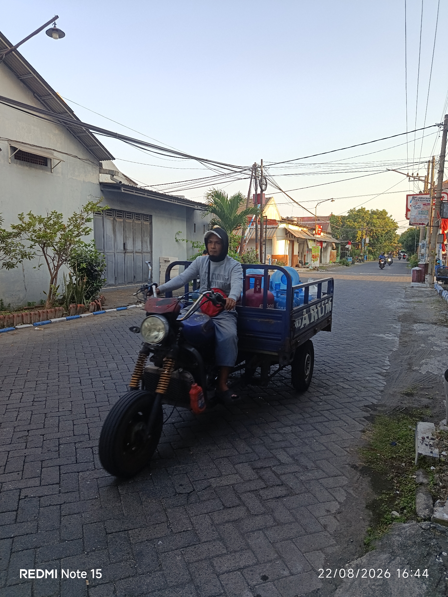 A man wearing a grey hoodie and a baseball cap is riding a three-wheeled motorcycle with a cargo bed. The cargo bed contains several large blue water jugs and a red propane tank. The motorcycle is dark blue and black with a visible radiator. The man is looking towards the camera. The motorcycle is on a cobblestone street in a residential area. Buildings with grey walls and traditional Indonesian architecture line the street. Power lines crisscross above the street. Palm trees and other vegetation are visible. In the background, other motorcycles are seen on the street. The sky is clear and blue. The image is timestamped "22/08/2026 16:44" and branded "REDMI Note 15." The location is identified as Keboansikep, Indonesia.