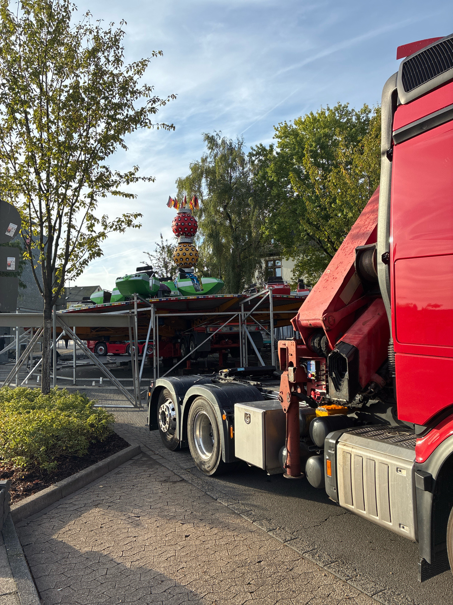 The image shows a red crane truck parked on a street in front of a temporary structure that appears to be part of a fairground ride. The ride itself is partially visible, with green and yellow pods and a tall, stacked sphere adorned with flags on top. The truck's crane is lowered and positioned over the ride. The scene is set outdoors during the daytime, with trees and buildings in the background. The street is paved with interlocking concrete blocks. The sky is partly cloudy. Based on accompanying metadata, the location is Radevormwald, Germany, on Kaiserstraße. There are no people visible in the image.
