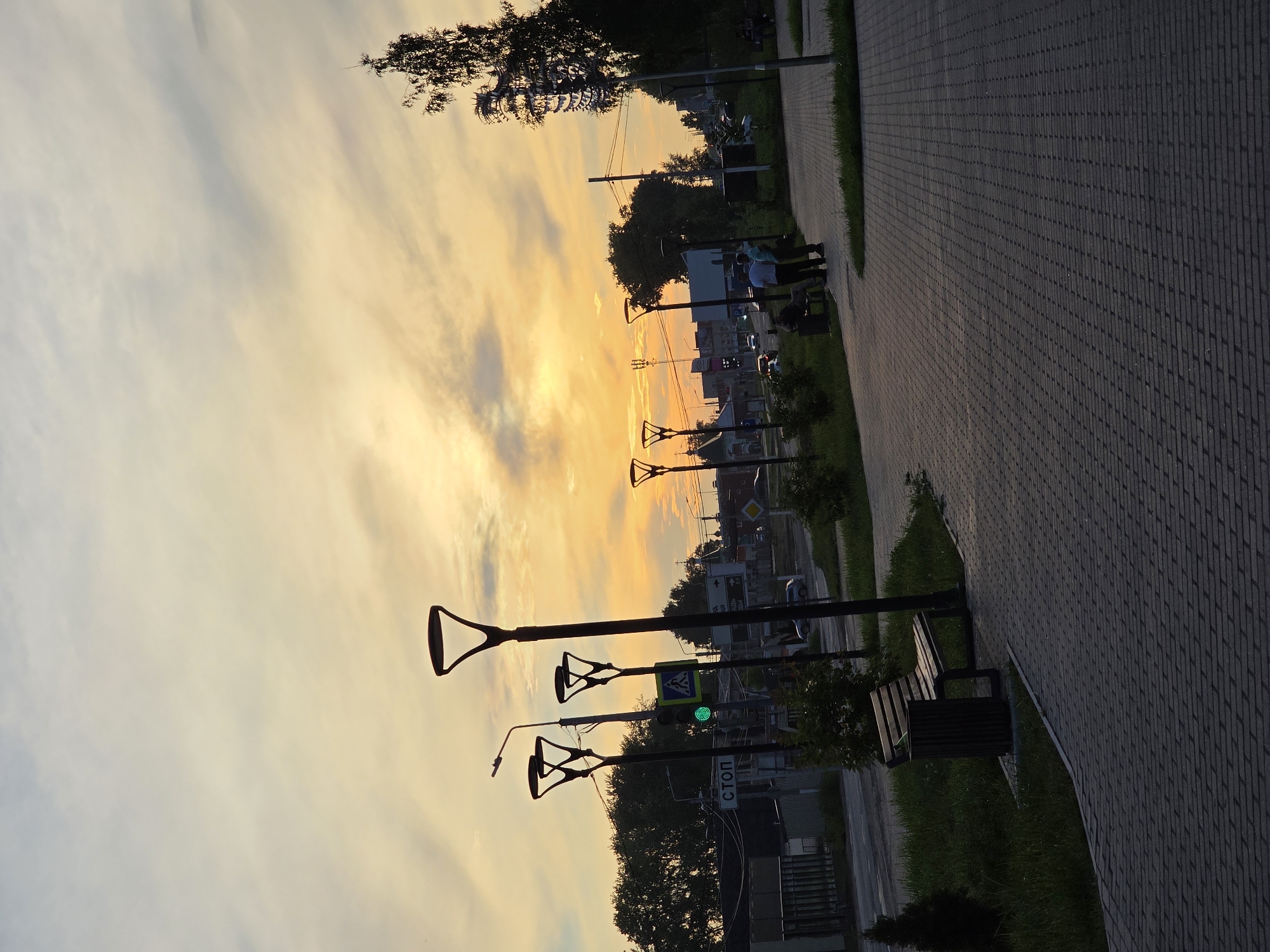 The image depicts a street scene at dusk in Chaikovsky, Russia. The sky is filled with a vibrant orange and yellow sunset, casting long shadows across the paved walkway and road. Several modern lampposts with angular designs line the sidewalk. A pedestrian crossing with a green traffic light is visible, along with a stop sign. In the background, trees and buildings form the urban landscape.
Several individuals are present in the scene. On the walkway, a group of three men are standing and conversing, one of whom appears to be seated on a bench. Further along the walkway, a solitary figure is seated on a bench near a trash receptacle.
The foreground is dominated by a grey brick-patterned walkway with grass and bushes bordering it. A small, conical evergreen tree is planted near the beginning of the walkway. The overall atmosphere is calm, suggesting the end of the day.