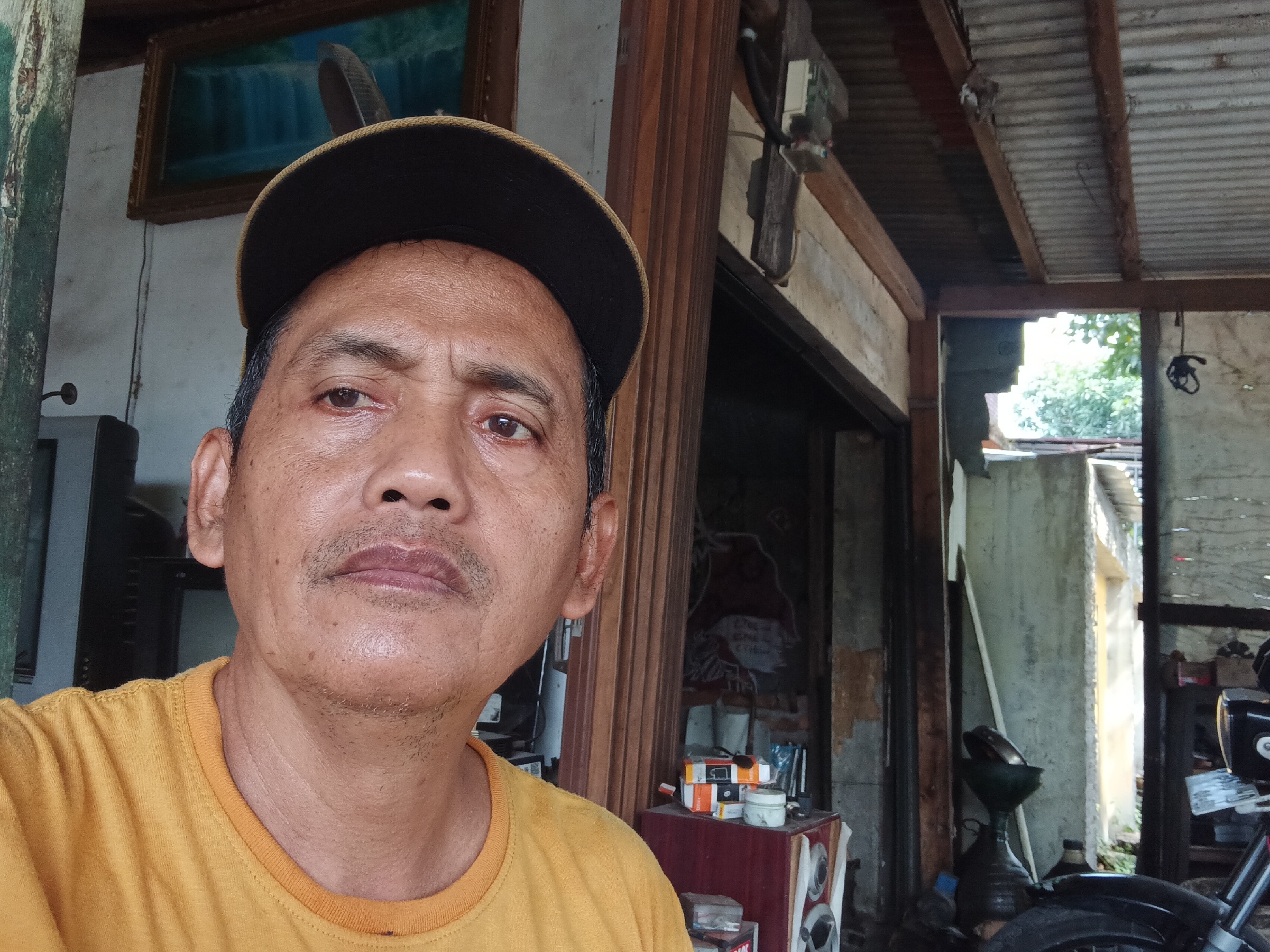 In Batursari, Indonesia, a close-up, selfie-style photograph captures a middle-aged man wearing an orange t-shirt and a dark baseball cap. He is positioned in the foreground, his face angled slightly to the right, with a neutral expression. Behind him, the scene depicts an outdoor, possibly workshop or yard area. Wooden posts and a corrugated metal roof are visible, suggesting a rustic or utilitarian structure. Various objects are scattered in the background, including what appears to be a motorcycle, shelves with items, and a framed painting on the wall, contributing to a sense of a lived-in, possibly cluttered space. The lighting is natural, indicating daytime.