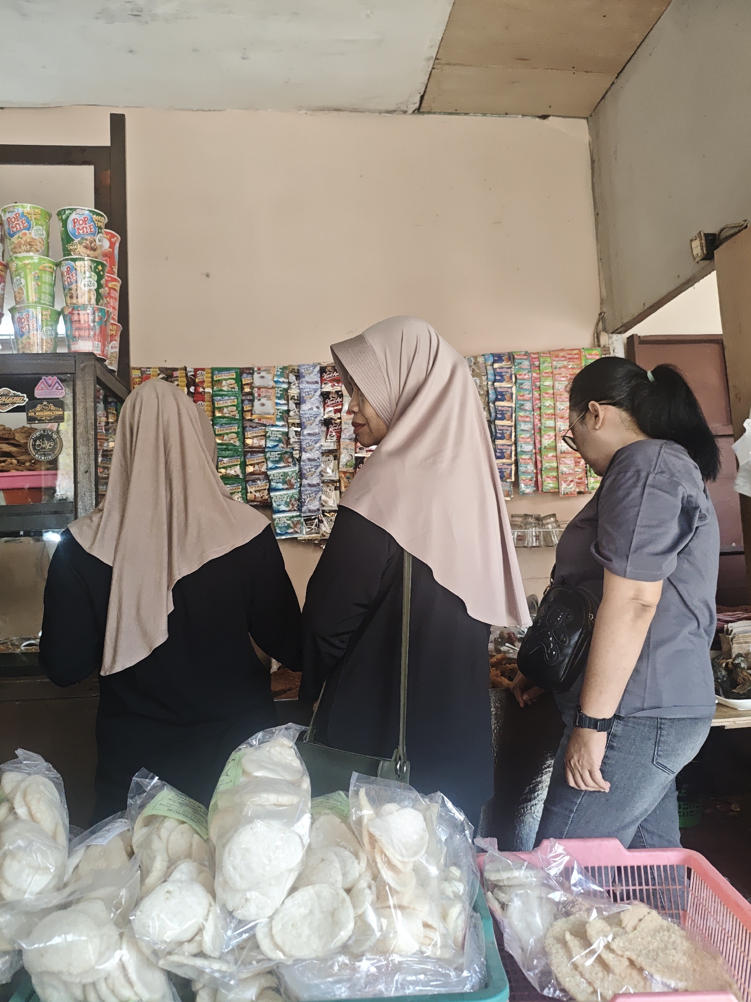 In the city of Lau, Indonesia, three women stand in a small shop. The woman on the left and the woman in the center are wearing light brown hijabs and dark tops, with their backs to the camera. The woman on the right is wearing a grey t-shirt and denim jeans, facing a wall lined with packets of snacks. In the foreground, several plastic bags of white, round snacks, possibly crackers, are arranged in green and pink plastic baskets. The shop appears to be a local convenience store or warung, with shelves displaying various food items.