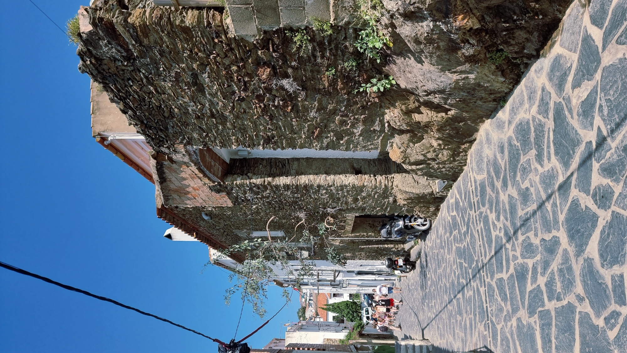 The image depicts a narrow, cobblestone street in Cadaqués, Spain. A steep, rocky hillside borders the right side of the street, with a rough stone wall constructed at its base. Several buildings with red-tiled roofs and whitewashed facades line the left side of the street. Some of these buildings appear to be old, with visible brickwork and rough stone construction elements.
A few people are visible walking down the street, seemingly engaged in casual conversation. Among them are men, women, and children, dressed in summer attire. A black motorcycle is parked on the left side of the street, partially obscured by a tree with sparse foliage. Another motorcycle is parked further down the street, also on the left.
The sky is a vibrant, clear blue, suggesting a sunny day. The lighting indicates it is likely midday, with distinct shadows cast across the cobblestone pavement by the buildings and a thin wire stretching across the scene. The overall impression is a charming, sun-drenched European village street. There is no visible text in the image.