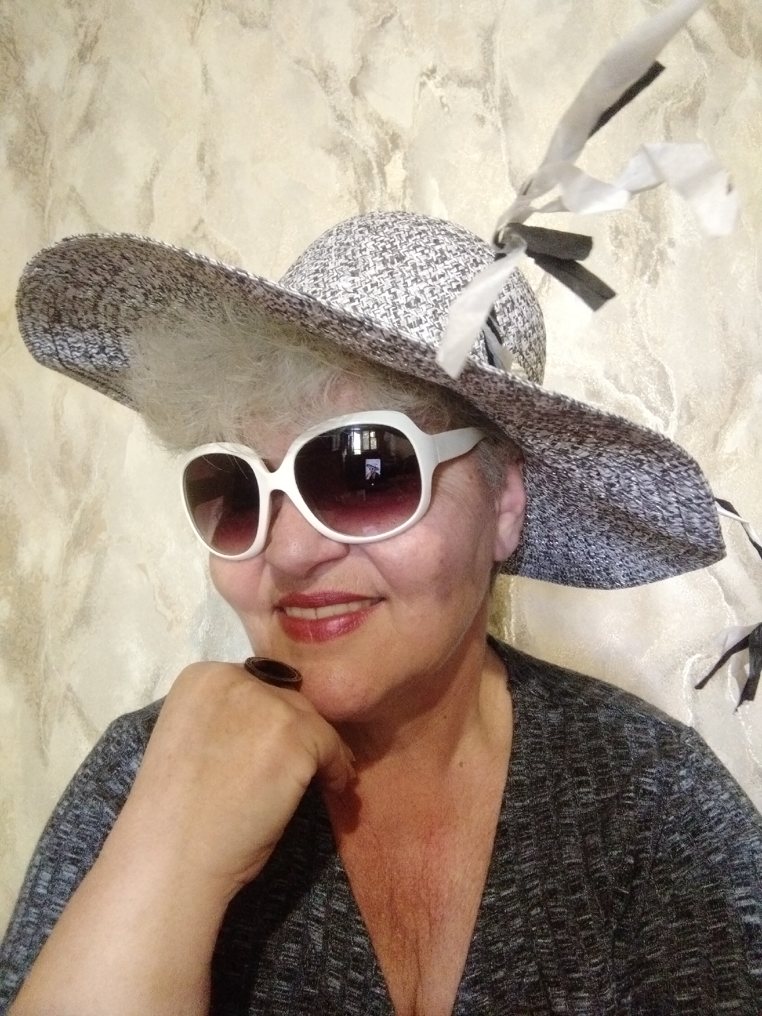 A woman with short, gray hair and white-rimmed sunglasses is wearing a wide-brimmed hat adorned with white and black ribbons. She is resting her chin on her fist, where she wears a large ring. She is wearing a dark gray, V-neck top. The background is a textured, light-colored wall. The location cannot be determined solely from this image, but based on the user's prompt, it is supposedly located in Horishni Plavni, Ukraine.