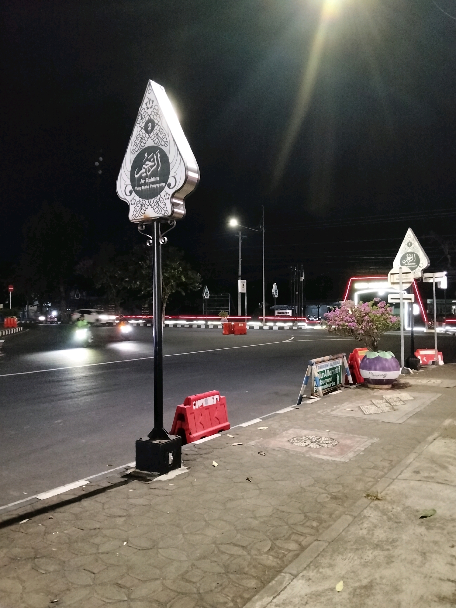An exterior night-time shot shows a street corner in Purworejo, Indonesia. A large, illuminated sign shaped like a Javanese wayang puppet is mounted on a pole. The sign features Arabic calligraphy and text in Indonesian. The street is wet and reflects the light from streetlights and vehicle headlights. Two motorcycles are visible on the road, their headlights illuminating the pavement. Red and white traffic barriers are placed along the side of the road. The sidewalk is paved with interlocking stones and a small flowering tree is planted in a pot. Further down the street, a brightly lit archway is visible, with more signs and traffic barriers. The overall scene suggests a public area at night, possibly a junction or intersection.