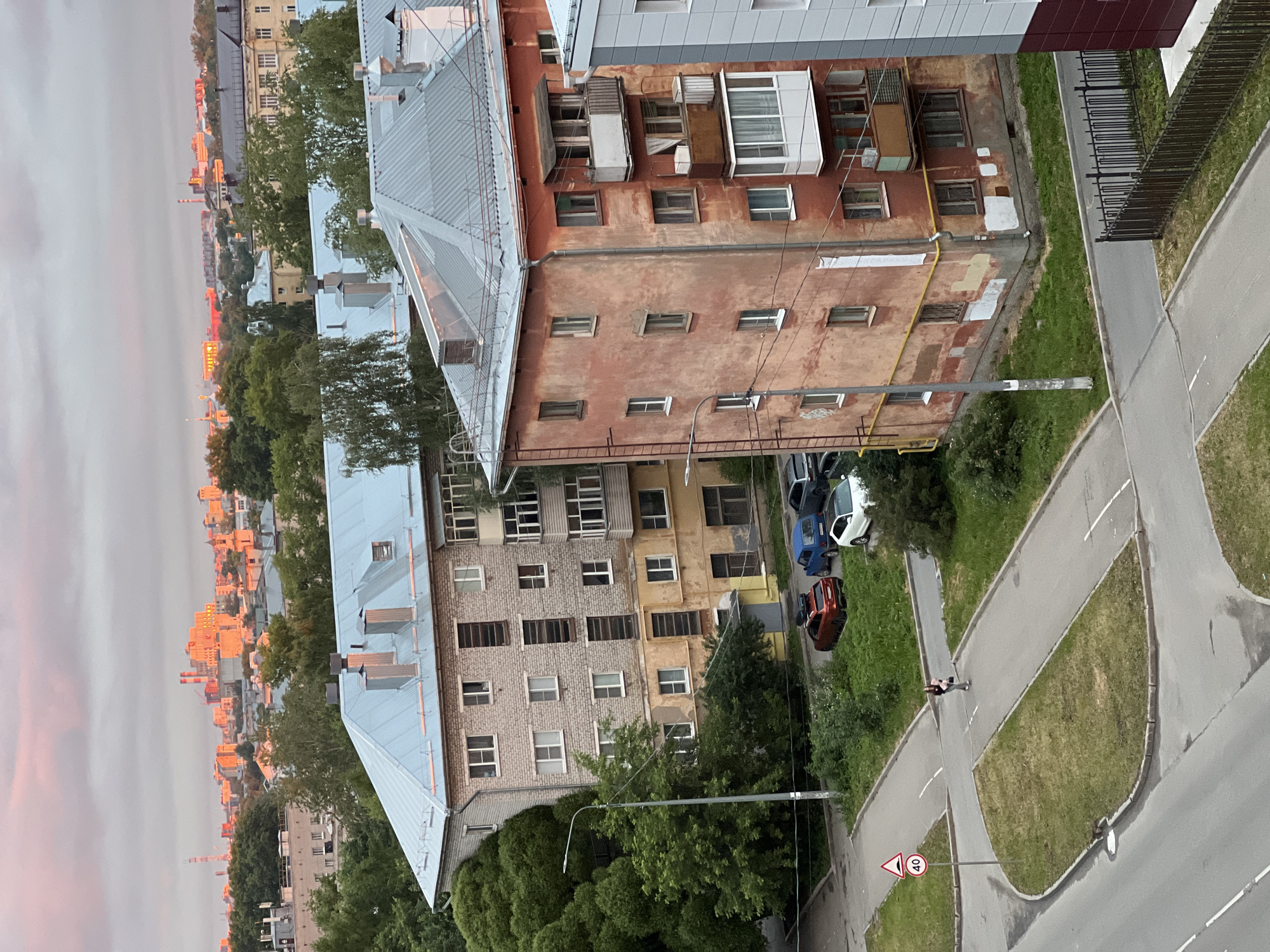 This aerial view captures a residential area in Vologda, Russia, at what appears to be dusk or dawn, with soft pink light illuminating the sky and distant buildings. Two multi-story apartment buildings dominate the foreground. The building on the left is clad in light brown brick with a light blue metal roof, featuring multiple windows and balconies. Adjacent to it is a larger, older building with a weathered, reddish-orange facade and a gray metal roof. This building exhibits signs of age, with peeling paint and some boarded-up windows.
In front of these buildings, a street intersection is visible with paved roads and grassy islands. Several parked cars are present: a red SUV with luggage on its roof, a blue hatchback, a dark sedan, and a white car. A single pedestrian, a person wearing dark clothing and carrying a bag, walks across the intersection. A speed limit sign indicating "40" is posted near the road. Lush green trees and bushes surround the buildings and the roadway, contributing to a suburban feel. In the distance, the Vologda cityscape is visible, with industrial buildings and other structures silhouetted against the colorful sky. A tall communication tower is also prominent in the background. The overall scene depicts a typical urban residential environment with signs of everyday life and the characteristic architecture of older Russian apartment blocks.