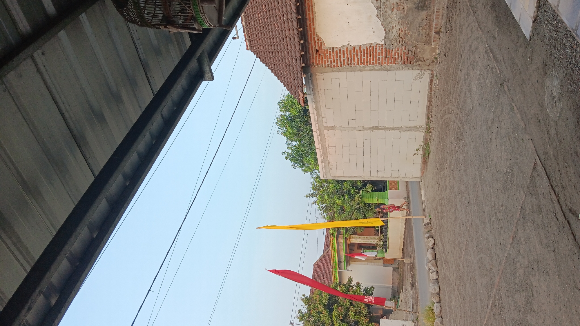 The image depicts an outdoor scene, likely in Sonobekel, Indonesia, under a clear sky. A corrugated metal roof structure extends into the foreground from the top of the frame. In the middle ground, a residential building with a red tile roof is visible, partially obscured by a wall constructed of white concrete blocks. A section of the wall shows exposed red brickwork.
To the left of the building, a person wearing a red and white patterned dress walks on a paved area. Two tall, thin flags are planted in the ground; one is red and the other is yellow. A smaller Indonesian flag, red and white, is attached to the front of the residential building. Green foliage from trees is visible behind the building and the wall.
The ground in the foreground is composed of dark, dusty soil with tire tracks visible. In the upper right corner, a wooden birdcage hangs from the roof structure. The overall impression is of a residential area during daylight hours, possibly in a village or a less urbanized part of the city.