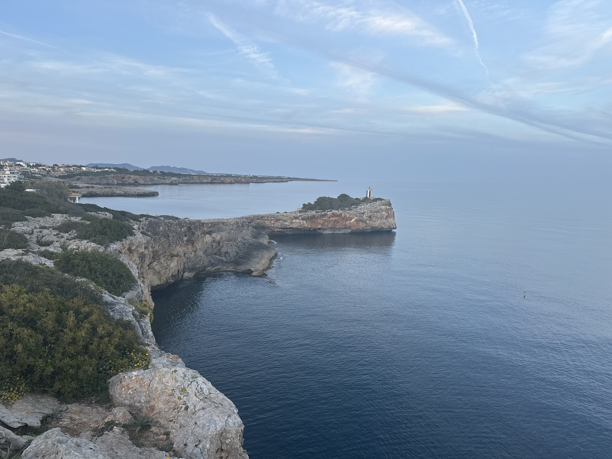 The image depicts a coastal landscape in Manacor, Spain, under a partly cloudy sky. Rocky cliffs with sparse vegetation dominate the foreground. To the right, a small promontory extends into the water, topped with a structure resembling a lighthouse or beacon. The water is relatively calm. Distant land is visible on the horizon.