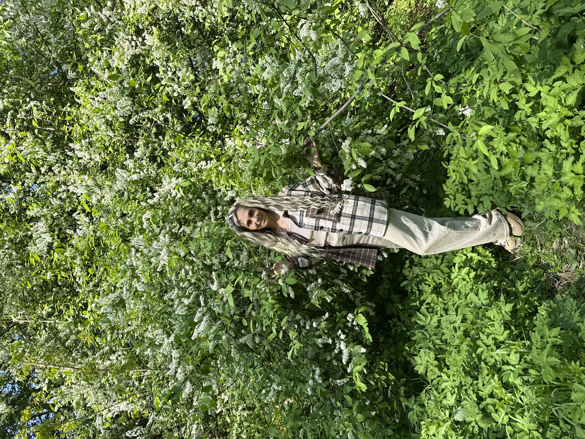 A blonde woman in a plaid jacket and beige pants stands amidst dense green foliage with white flowers. The location is the city of Rusanovo, Russia.