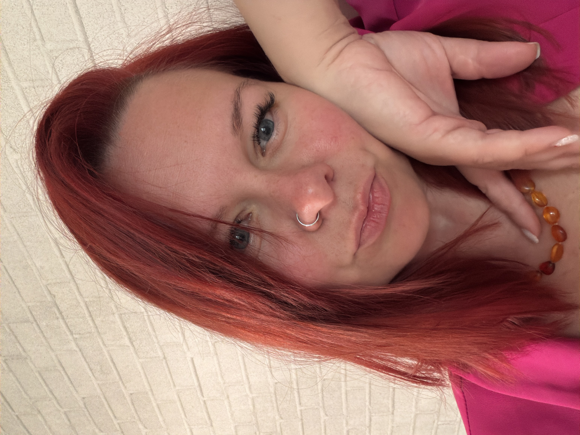 A close-up, selfie-style shot features a fair-skinned woman with red hair. She has blue eyes and a silver nose ring in her left nostril. Her right hand is partially visible, resting near her face. She is wearing a pink top and an amber-colored beaded necklace is partly visible. The background is a textured white wall. There is no information to suggest the location is in Chaikovsky, Russia.