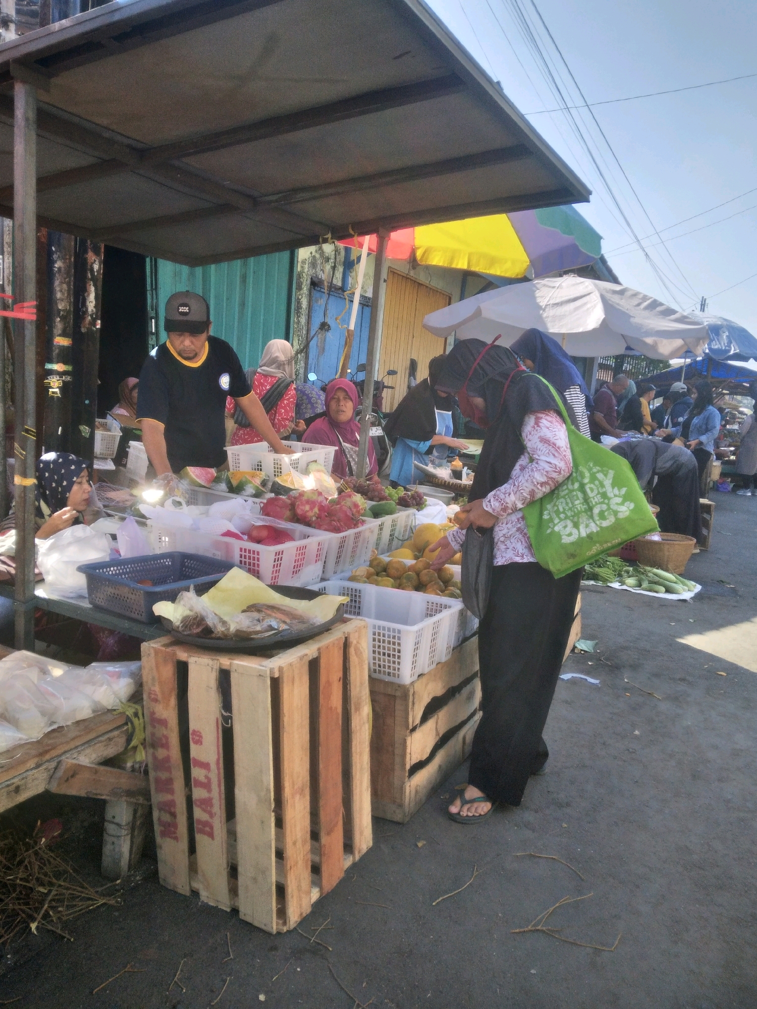 In a market stall in Sokaraja, Indonesia, a vendor wearing a black t-shirt and a dark baseball cap stands behind a table laden with fruit. The fruit is displayed in white plastic crates, including dragon fruit, watermelons, and other produce. To the right of the vendor, a woman in a patterned pink and white shirt and a dark headscarf, holding a green reusable shopping bag, is reaching for fruit. Another woman in a pink headscarf is visible behind the fruit display. In the foreground, a wooden crate with the words "MARKET BALI" is positioned on the ground. The market stall is covered by a corrugated metal roof. The ground is paved with asphalt, and dried branches and twigs are scattered across it. Several other people and market stalls are visible in the background. The sky is clear and blue, suggesting a sunny day.