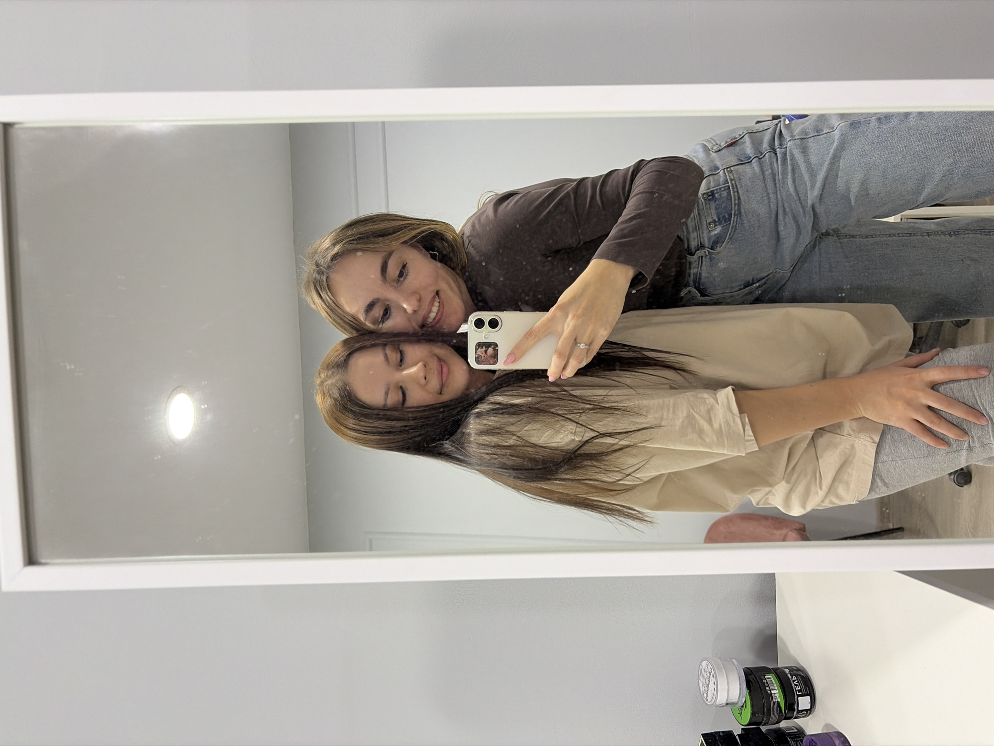 Two women are taking a selfie in a bathroom mirror. One woman has brown hair and is wearing a brown long-sleeved shirt and jeans. The other woman has long brown hair and is wearing a beige top and grey pants. The mirror is white-framed, and reflected in it are the women and the bathroom interior. There is a white countertop with various haircare products in the foreground. The bathroom has white walls. There is no evidence to suggest that this image was taken in the city of Izhevsk, Russia.