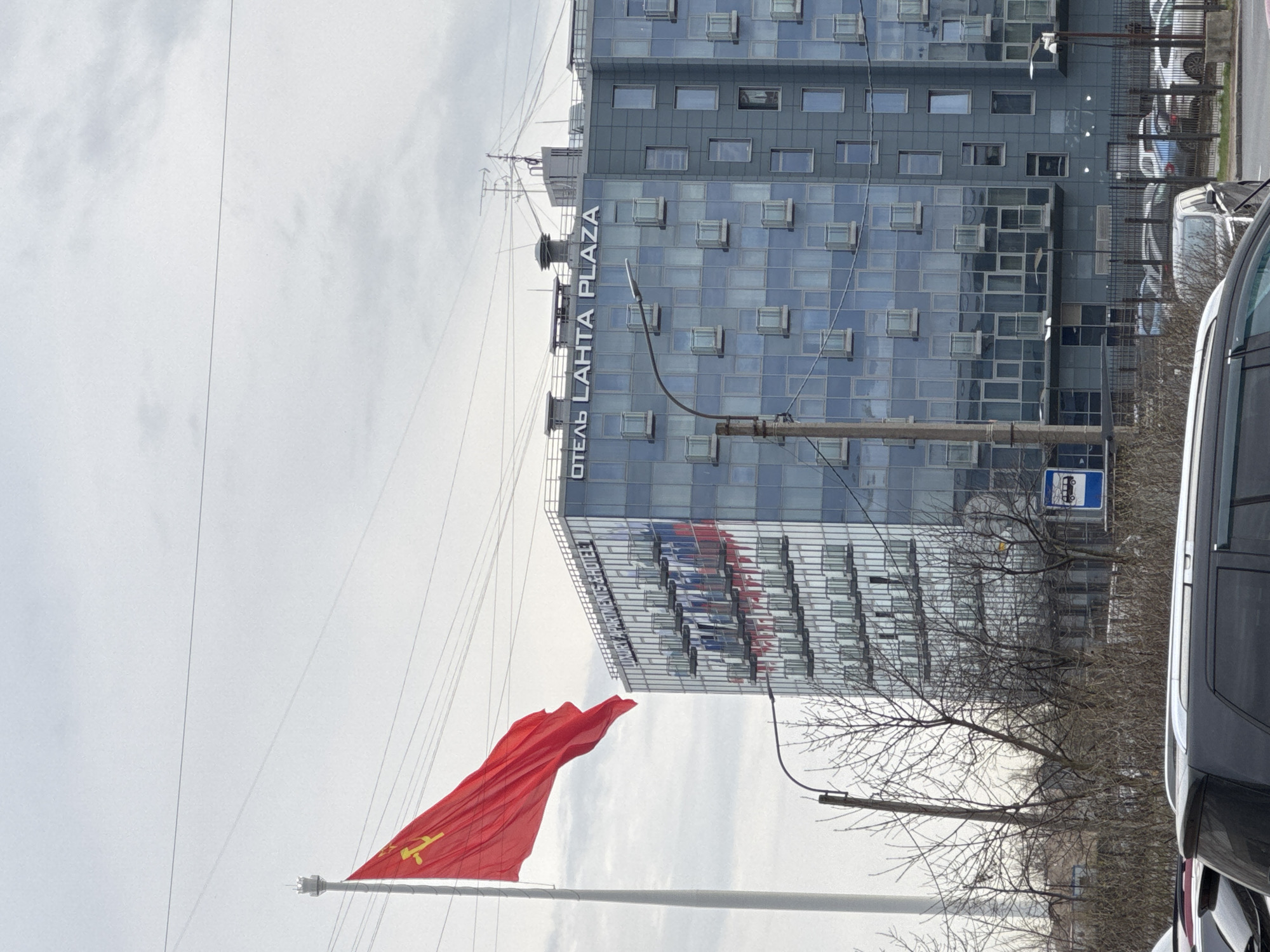 The image captures a red flag emblazoned with a yellow hammer and sickle emblem, the symbol of communism, waving prominently in front of the Oтель Lahta Plaza building in Saint Petersburg, Russia. The building is a modern structure with numerous windows and a sign on its facade. In the immediate foreground, there's a partial view of a dark-colored vehicle, suggesting the photograph was taken from a roadside perspective. Bare branches of a tree are visible, interspersed with street furniture such as lamp posts and a bus stop sign. The sky is overcast, providing a muted backdrop.