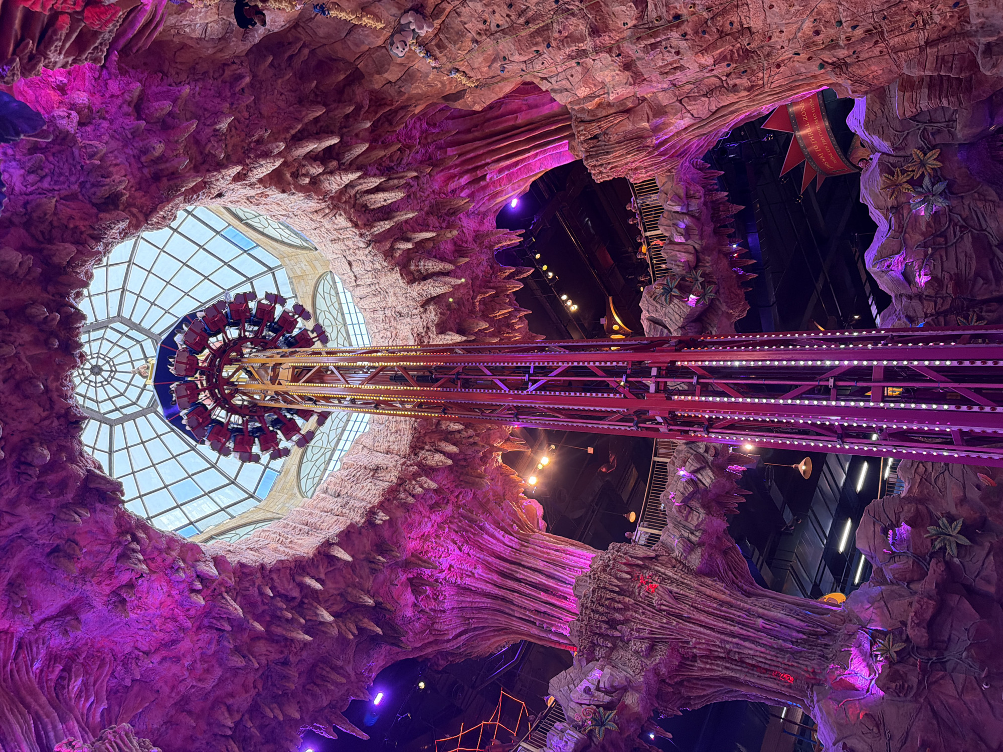 This is a photograph taken from the interior of Sun World Danang Wonders in Da Nang, Vietnam, looking upwards at an amusement park ride. The ride is a drop tower, with a central red structure featuring vertical lines of purple LED lights. A circular platform at the top of the tower carries passengers in red seats, and the platform is in motion. Surrounding the ride are massive, artificial rock formations designed to resemble a cave or grotto, illuminated with vibrant pink and purple lighting. These formations have stalactite-like protrusions hanging from their ceilings and textured rock faces. Above the ride and rock formations, a large, segmented glass ceiling is visible, allowing natural light to filter in. In the background, the darker interior of the amusement park is visible, with other structures, railings, and people on higher levels. A banner with red text on a yellow background is also visible in the middle distance. The overall impression is one of an indoor amusement park with a fantasy or adventure theme.