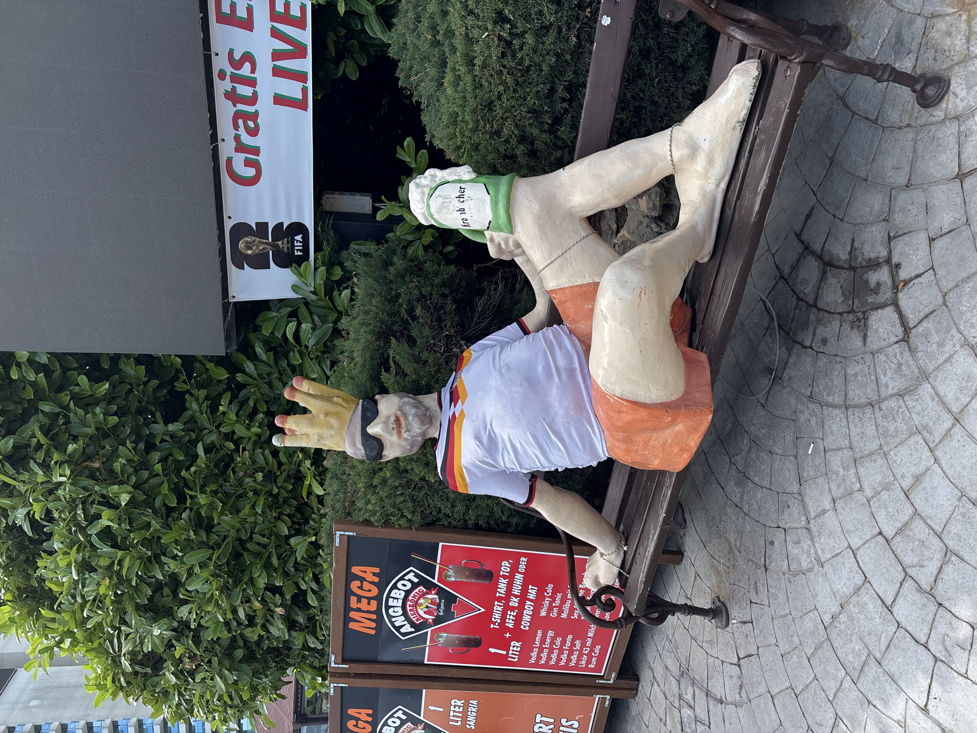 This media file captures a quirky roadside scene in Varna, Bulgaria, featuring a life-sized, somewhat weathered statue of a man lounging on a park bench. The statue, dressed in a German-themed sports jersey and orange shorts, wears a golden crown and sunglasses, giving him a playful, king-like demeanor. He appears to be enjoying a beverage, with one hand reaching towards a sign.
The bench itself is made of dark wood and metal, showing signs of age. The ground beneath is paved with circular stone patterns, and a variety of greenery, including lush bushes and trees, forms the backdrop. To the left of the statue, a large banner displays the text "Gratis E LIVE" and the FIFA logo, suggesting the presence of a sports-related venue or event, possibly a bar or restaurant advertising free live broadcasts.
Below the statue, a prominent sign advertises "MEGA ANGEBOT" (Mega Offer) from "BIERKONIG," featuring drinks like Sangria, Vodka Lemon, Whisky Cola, and Gin Tonic, along with a "T-SHIRT, TANK TOP, + AFFE, BK HUHN ODER COWBOY HAT" package. This indicates the location is likely a bar or pub offering special deals. Another smaller sign below suggests "1 LITER SANGRIA."
The statue's playful pose and the vibrant advertising suggest a lively and possibly tourist-oriented atmosphere. The lighting indicates it's daytime, and the abundant greenery suggests fair weather. The overall impression is one of a casual, fun-loving establishment aiming to attract customers with eye-catching displays and promotional offers.