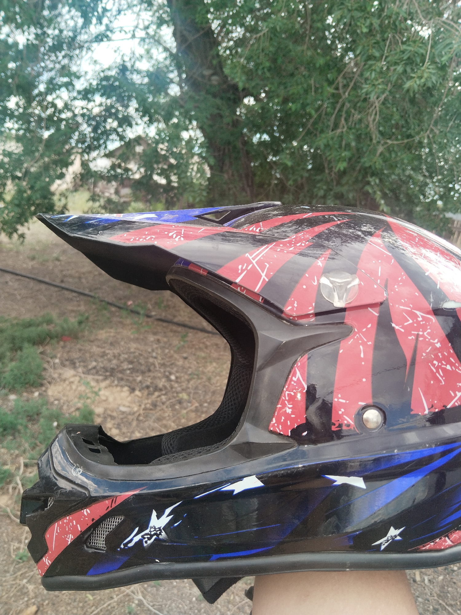 This is a close-up, medium shot of a black and red motocross helmet with blue accents and white star graphics. The helmet is angled to the right, showing its profile. The sun visor is prominent and angled upwards. The interior padding is visible through the opening. The helmet has a rugged, worn appearance with some scuffs and scratches. In the background, there is out-of-focus foliage from trees and dry, earthy ground, suggesting an outdoor setting in a natural environment. The lighting indicates it is daytime. The image was taken in Ақадыр, Kazakhstan. No text is visible.