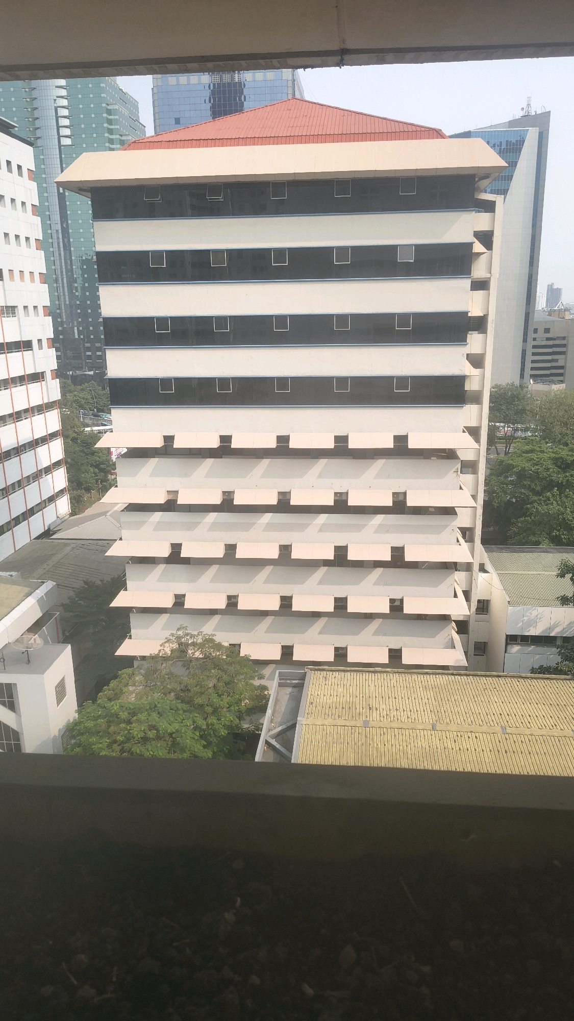 The image is an outdoor, eye-level, full shot of a multi-story building with a red roof, situated in a city environment. The building is predominantly cream-colored with horizontal black stripes and numerous rectangular windows. Several floors feature awnings or sunshades. To the left, another taller building with white vertical elements and numerous windows is partially visible. In the background, other city buildings, including a modern skyscraper with a blue-tinted glass facade, are present. Trees and greenery are visible around the buildings. The foreground is dominated by a dark, textured surface, possibly gravel or soil, and a concrete ledge. The overall impression is an urban landscape in daylight, likely South Jakarta, Indonesia, given the context of other images from this set. There are no people or animals visible in the frame.