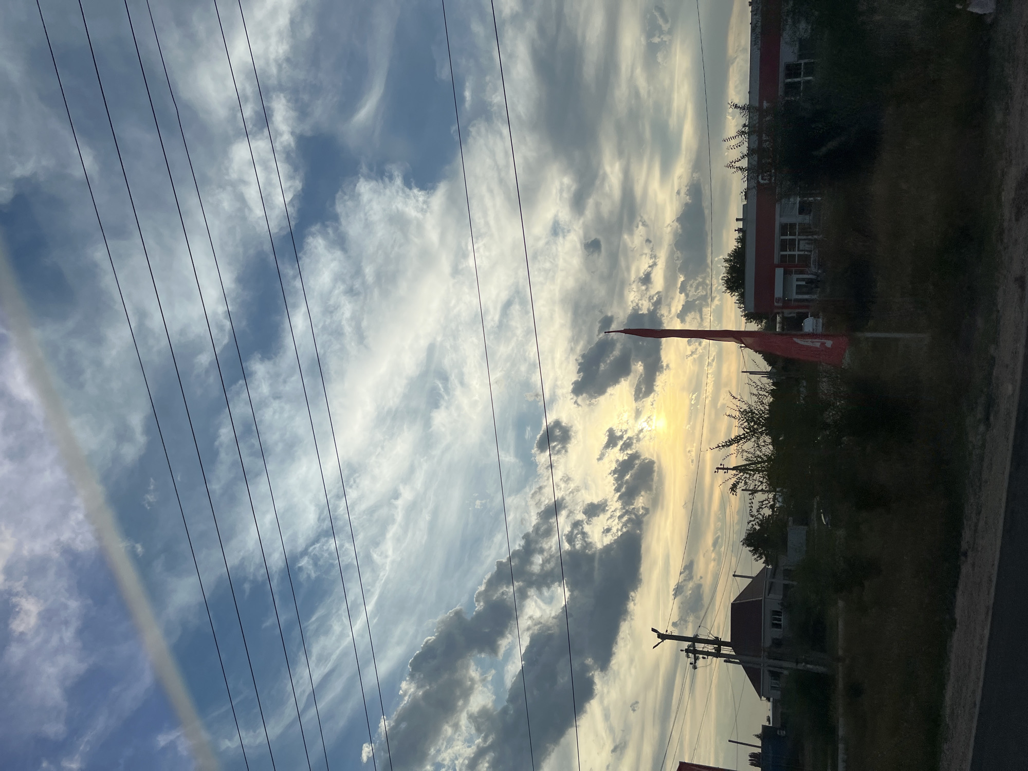 The image captures a wide-angle view of a rural roadside scene in Shumeyka, Russia, under a dynamic sky. The sky is a prominent feature, filled with a mix of cirrus and cumulus clouds, illuminated by a soft, hazy sunset. A prominent contrail stretches diagonally across the upper portion of the frame, adding a linear element to the cloudy expanse. Several horizontal power lines crisscross the sky, obscuring parts of the cloud formations.
In the midground, a single-story building with a red-orange trim and white facade is visible, suggesting a commercial establishment. Adjacent to it are dense green trees and bushes. To the left of this building, a house with a dark red roof is partially visible, surrounded by greenery. A utility pole with power lines stands between the house and the road. In the foreground, dry grass and weeds line a paved road or pathway. A tall, bright red flag on a pole stands prominently in the middle ground, to the left of the commercial building, and its shadow falls to the right. The overall impression is one of a quiet, somewhat overcast evening in a semi-developed rural area. There are no people visible in the image.