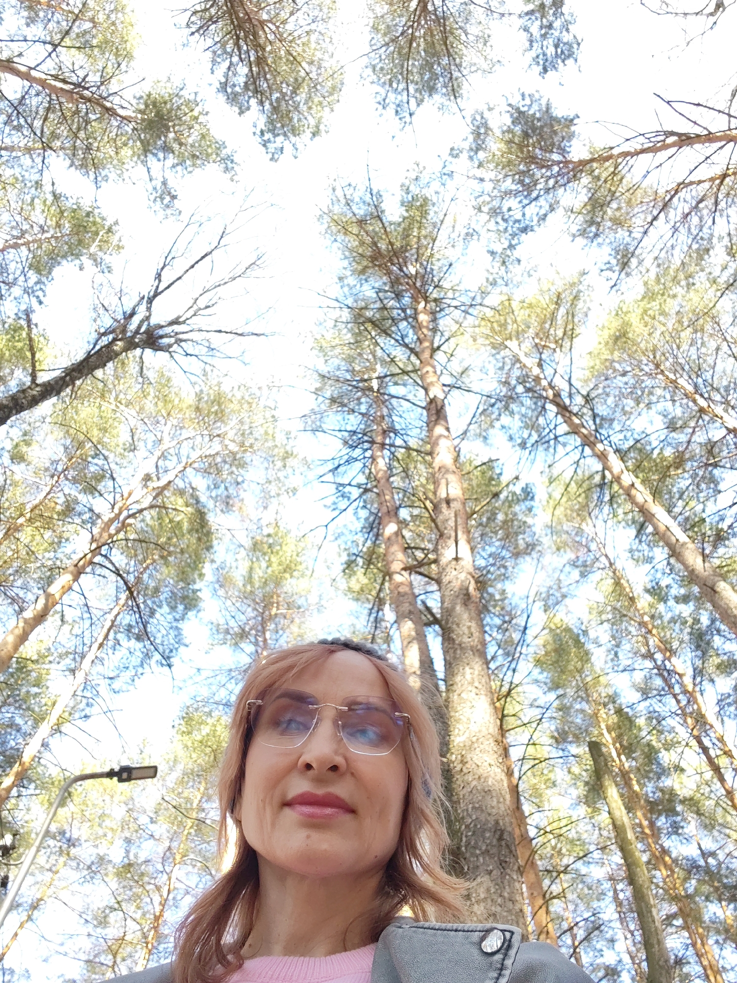 A low-angle, full-color photograph features a fair-skinned woman wearing glasses, positioned in the lower-center of the frame. She appears to be looking directly at the camera. Above her, the sky is visible through a dense canopy of tall trees. To the left of the woman is a lamppost. The photograph was taken in the city of Chaikovsky, Russia.