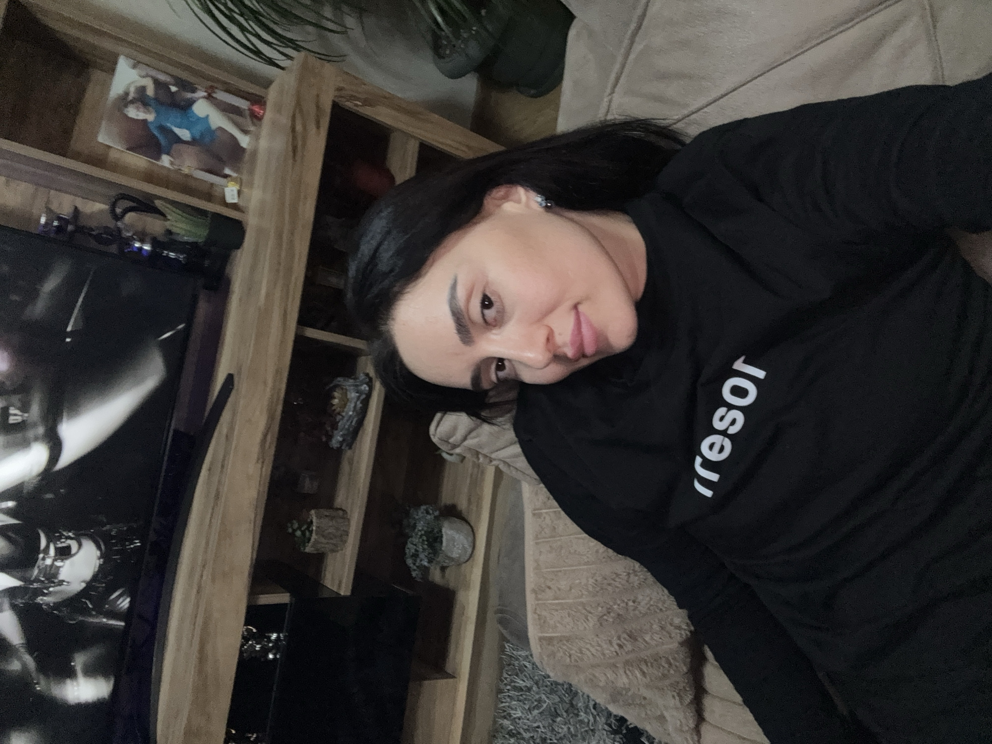 A woman with dark hair is lying on a light-colored, textured couch. She is wearing a black long-sleeved shirt with the word "resor" printed on it. To the left of the couch, there is a tall wooden shelving unit with various items, including a picture of a person in a blue leotard, plants, and decorative objects. A large black television or monitor is also visible. The location cannot be determined based on visual information.