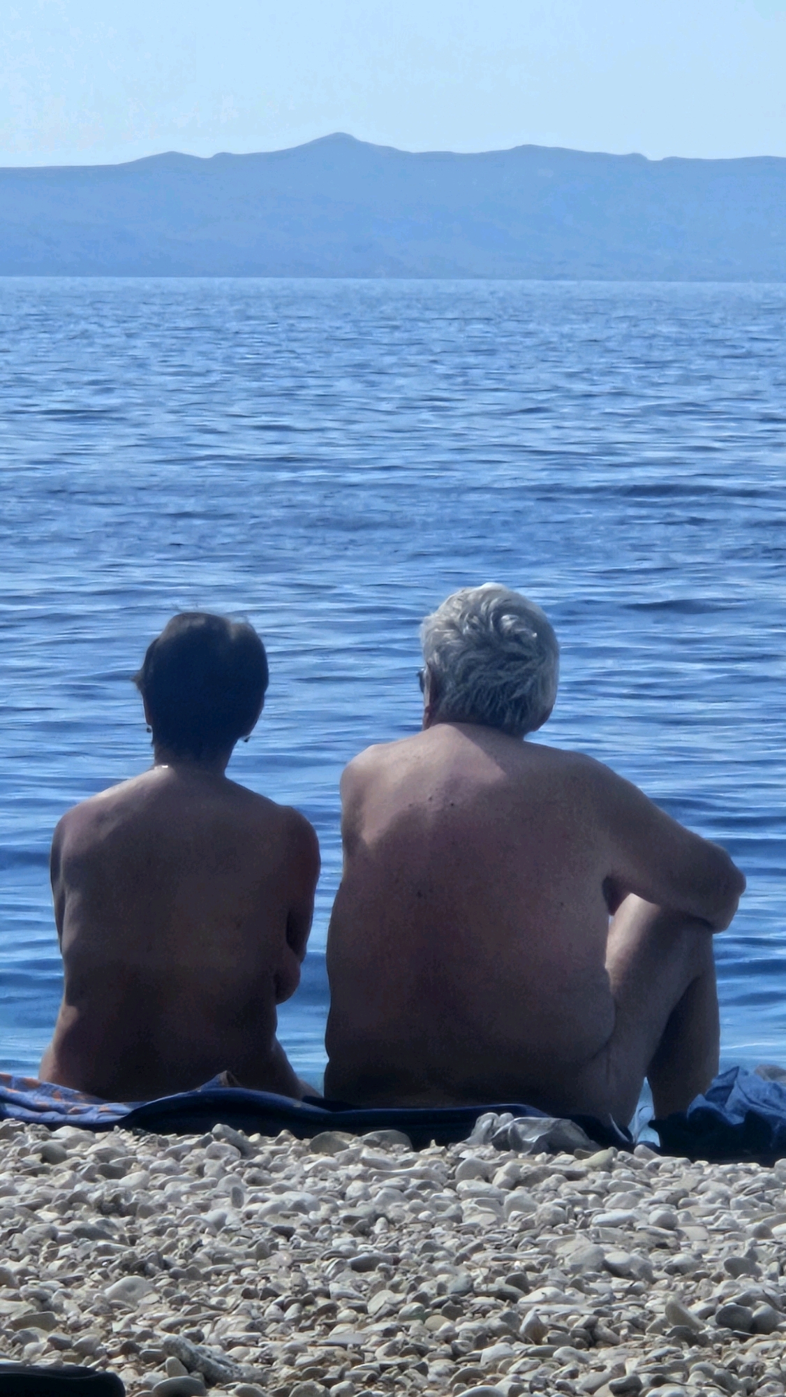 Two individuals are seated on a pebble beach, facing a body of blue water, with a distant mountain range visible on the horizon. The individuals, one with dark hair and the other with grey hair, appear to be an older couple. They are both unclothed and sitting with their backs to the camera. A blue towel is positioned beneath them, and a blue garment is visible to the right. The scene is set in Općina Podgora, Croatia.