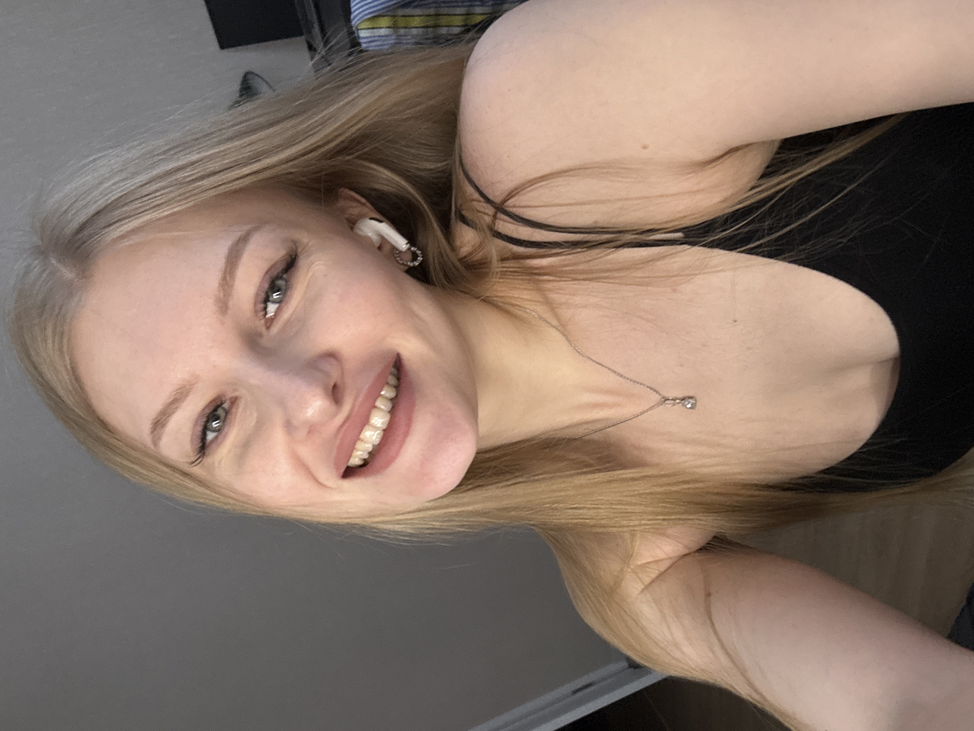 The image features a young woman with long, straight blonde hair, appearing to be taken as a selfie indoors. She is smiling broadly and looking directly at the camera, with light-colored eyes (grey or green) and well-defined eyebrows. Her smile reveals her upper teeth and creates natural crinkles around her eyes, conveying a cheerful and friendly demeanor.
She is wearing a black camisole or tank top, which is partially visible. A delicate silver chain necklace with a small, clear pendant hangs around her neck. A white wireless earbud, resembling an AirPod, is visible in her right ear. Her blonde hair is parted on the side and flows around her shoulders.
The background is minimalist, consisting mainly of a plain light grey wall, suggesting an indoor setting, possibly in a room in Perm, Russia. A hint of a striped pattern, potentially from bedding or a pillow, is visible behind her head in the upper right portion of the frame, along with some indistinct dark areas. The lighting is soft and even. There is no visible text in the image. The photograph is oriented diagonally, adding to its casual, candid feel.