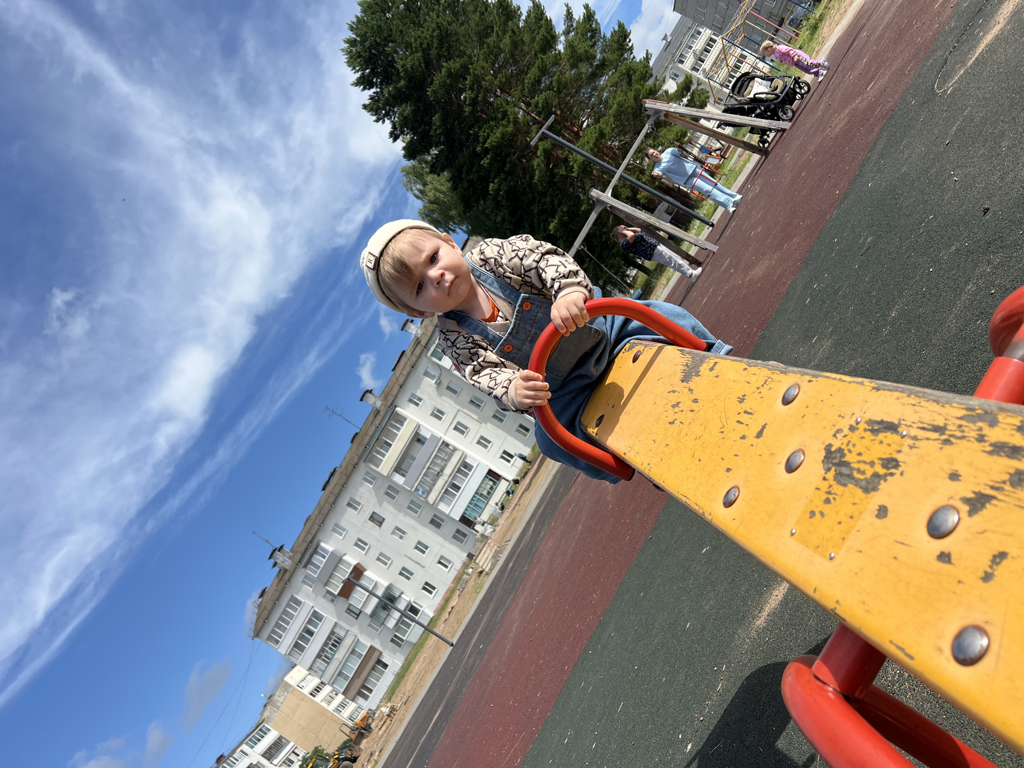A young child, possibly a toddler, is seated on a bright yellow seesaw with red handles. The child wears a white cap with a logo, a patterned long-sleeved shirt, and denim overalls. The child is looking directly at the camera with a neutral expression. In the background, there is a playground with swings and other playground equipment. Several adults and a young girl are visible in the middle ground and background. Beyond the playground, there are multi-story apartment buildings, characteristic of Noviy, Russia, under a partly cloudy blue sky. The ground surface of the playground is a combination of red and dark green safety surfacing. A large evergreen tree stands prominently to the right of the child. The scene appears to be a typical day at a neighborhood playground.