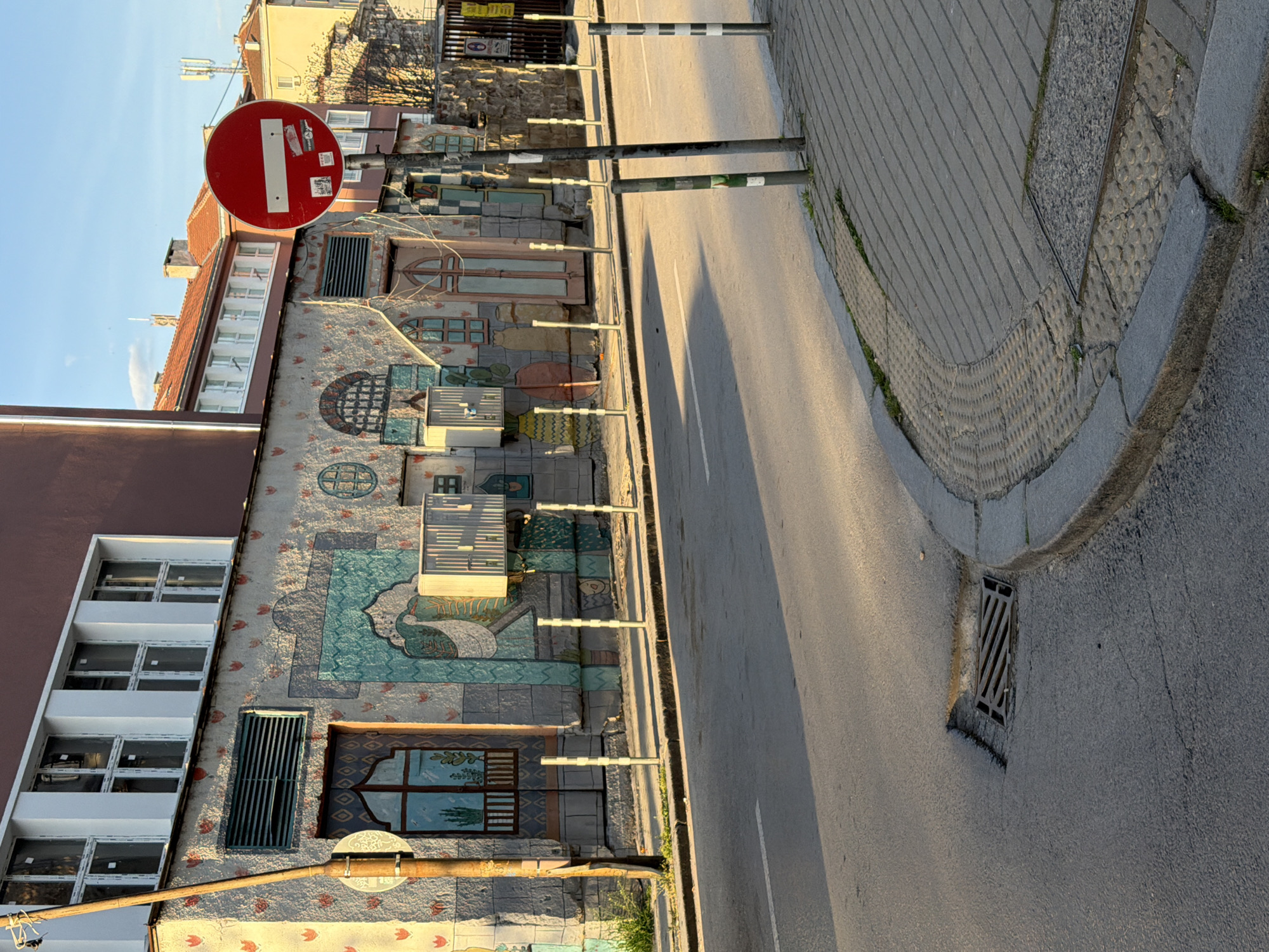 An outdoor, eye-level, upright shot captures a vibrant urban street scene during late afternoon. The dominant feature is a multi-story building on the left, adorned with an extensive, colorful mosaic-like mural covering its facade. This artwork depicts various stylized patterns, abstract shapes, and what appears to be a large bird figure. Rectangular and arched elements resembling windows are integrated into the design, some with blue and green hues. To the far left, a more modern building with multiple white-framed windows is partially visible.
To the right of the art-covered building, a narrow asphalt road stretches into the distance, marked by a single white dividing line. A metal drain grate is visible on the road surface in the foreground. Adjacent to the road is a paved sidewalk, featuring sections of tactile paving with raised dots and parallel lines, particularly visible on a curved corner. A series of thin, dark bollards or poles with white reflective stripes line the edge between the building and the road.
Prominently placed above the art-covered building, a red circular "No Entry" traffic sign with a horizontal white bar is attached to a taller dark pole. Small, unidentifiable stickers are affixed to the sign, but no readable text is visible. Another pole with a circular, frosted glass light fixture is visible lower down on the left. Long shadows are cast across the road and pavement by the poles, indicating a low sun angle, consistent with late afternoon or early evening. The sky above is clear and light blue. The overall atmosphere is peaceful, highlighting the unique architectural decoration.