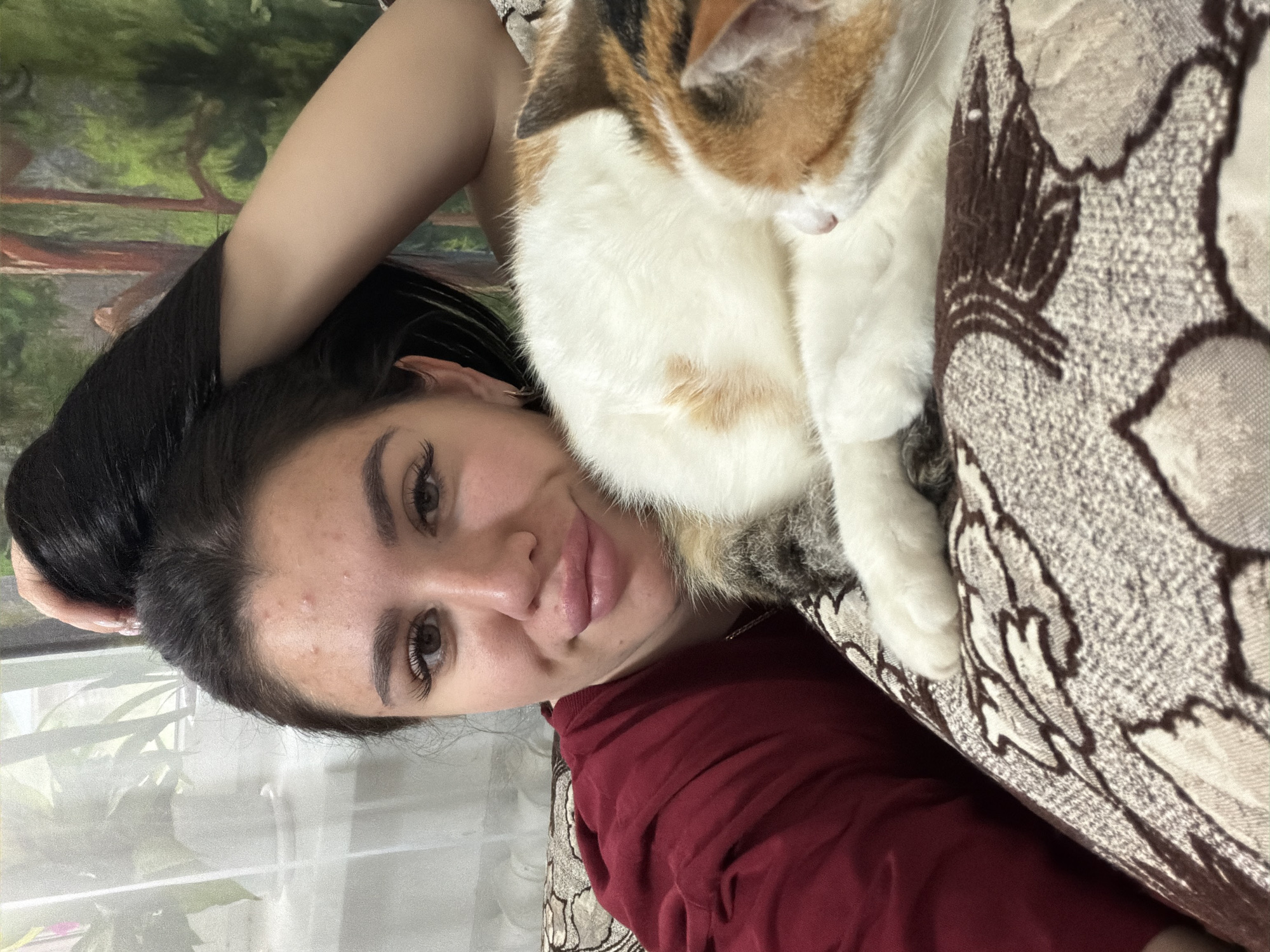 A young woman with dark hair pulled back is taking a selfie while resting her arm on a pillow. A calico cat is curled up beside her. The woman is wearing a red top and has full lips. The background appears to be an indoor setting with a window and a tapestry depicting trees.