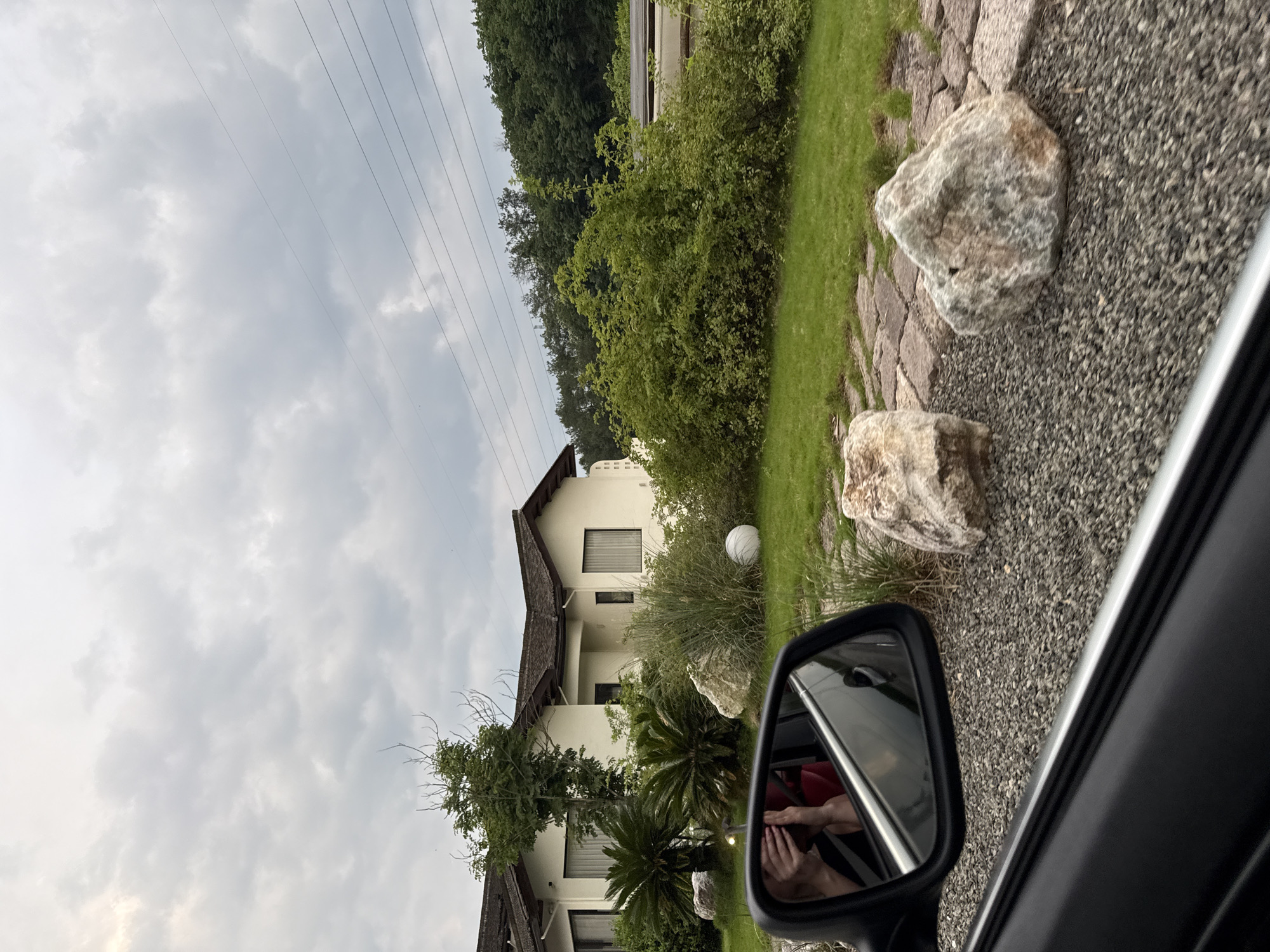 The image shows a view from inside a car, likely a passenger side window. Reflected in the side mirror are a person's hands holding a cell phone. Outside the car, the scene is dominated by a two-story, white stucco building with a dark brown roof. The building is surrounded by landscaping, including various green plants, bushes, and trees. There are large, light-colored rocks in the foreground near a gravel area. Overhead are power lines against a cloudy sky. While the city, Jinhua, is provided, there are no definitive elements within the image to confirm the exact location.