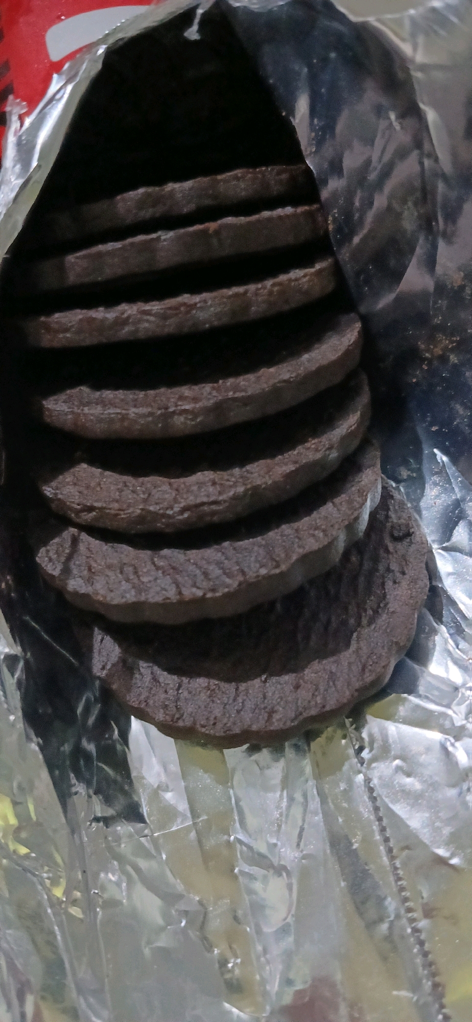 A stack of dark brown, round cookies is piled within a silver foil wrapper. The wrapper is crumpled and partially open, revealing the cookies inside. The cookies appear to be of uniform size and thickness, with a slightly rough texture. The lighting is dim, casting shadows on the cookies and the wrapper. No people or specific location details within Sutawinangun, Indonesia are visible in this image.