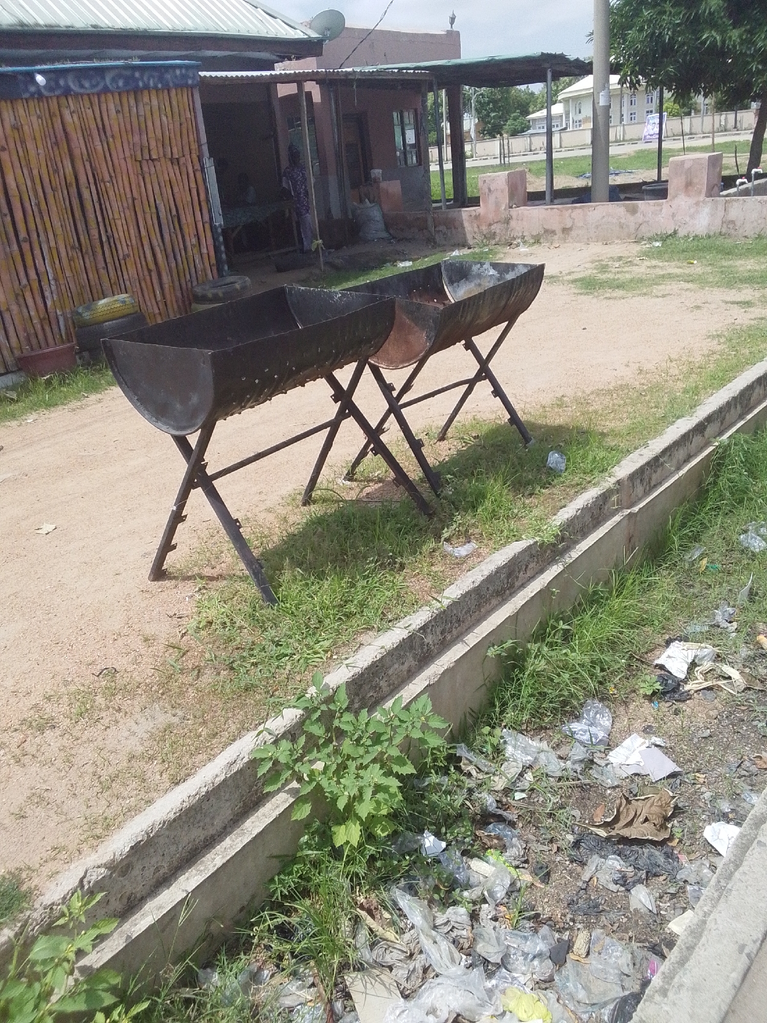 This is a medium-angle, outdoor shot of a scene in Jalingo, Nigeria. In the foreground, two black metal barbecue grills are set up on X-shaped stands. The grills appear to be made from cut metal barrels. To the right of the grills, a concrete drainage ditch runs along the edge of a sandy area. This ditch is filled with grass and littered with various types of trash, including plastic bags, paper, and other debris. In the background, a building with a corrugated metal roof and a bamboo-covered section is visible. There is a person standing near the entrance of the building, partially obscured. The ground is a mix of sand and sparse grass. Beyond the building, a street with a few cars and modern buildings can be seen in the distance. The sky is overcast.