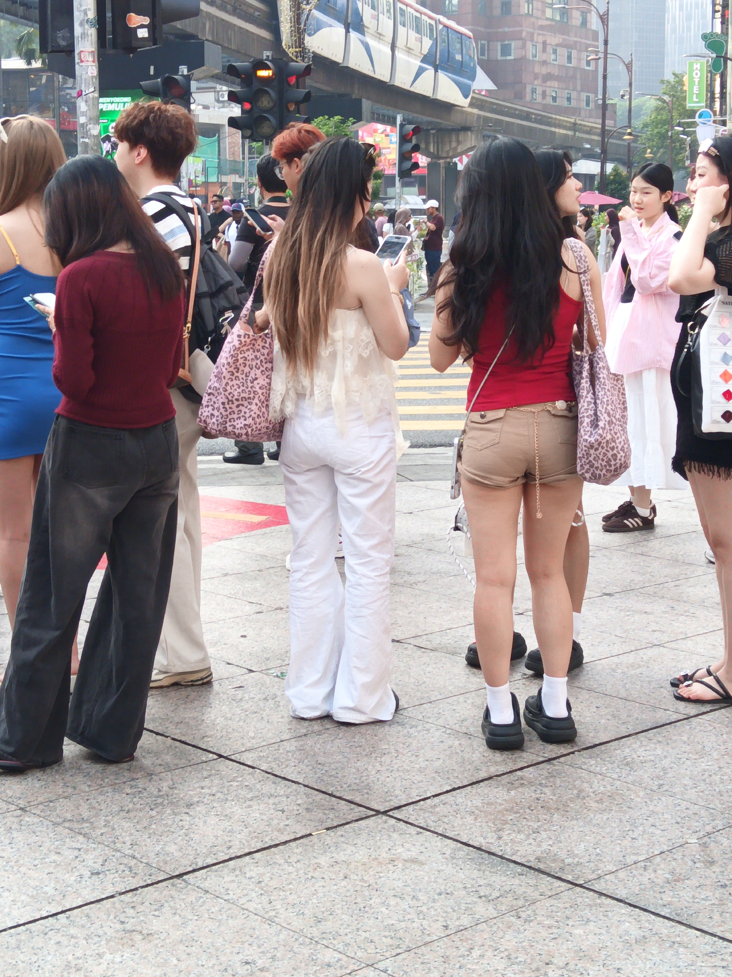 In Kuala Lumpur, Malaysia, on a paved pedestrian walkway with intersecting dark lines, a group of young women stand near a busy street intersection. A monorail train passes overhead on an elevated track. Traffic lights, including one displaying a red signal, are visible. The women, dressed in various casual outfits, are positioned around a street crossing. One woman on the left wears a blue dress and dark grey wide-leg pants. Next to her, a woman in a maroon top and grey pants holds a pink leopard-print bag. Another woman in the center wears a white lace top and white wide-leg pants. To her right, a woman in a red tank top and tan shorts holds a patterned bag. The background shows buildings and other pedestrians.