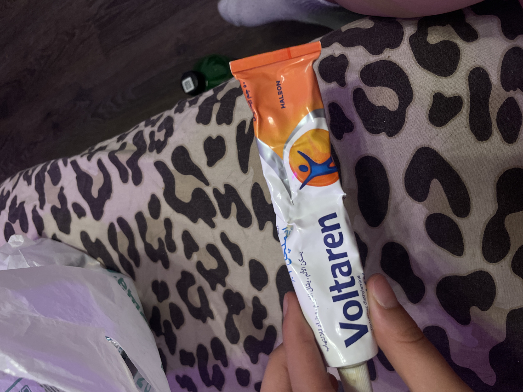 A tube of Voltaren, an anti-inflammatory gel, is being held by a person's hand against a leopard-print fabric. The tube is white and orange, with the brand name "Voltaren" prominently displayed in blue lettering. The text on the tube is in Arabic and appears to describe the product's uses. The background is a dark wood-like surface. A white plastic bag is partially visible in the upper left corner of the image. The image was taken in Vaganovo, Russia.