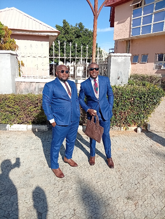 Two men in matching blue suits and sunglasses stand on a paved surface next to hedges and buildings. The man on the right carries a brown leather bag. Both wear brown leather shoes and ties with distinct patterns. The setting appears to be outdoors on a sunny day, as evidenced by the distinct shadows cast on the ground. A low metal fence is visible behind them.