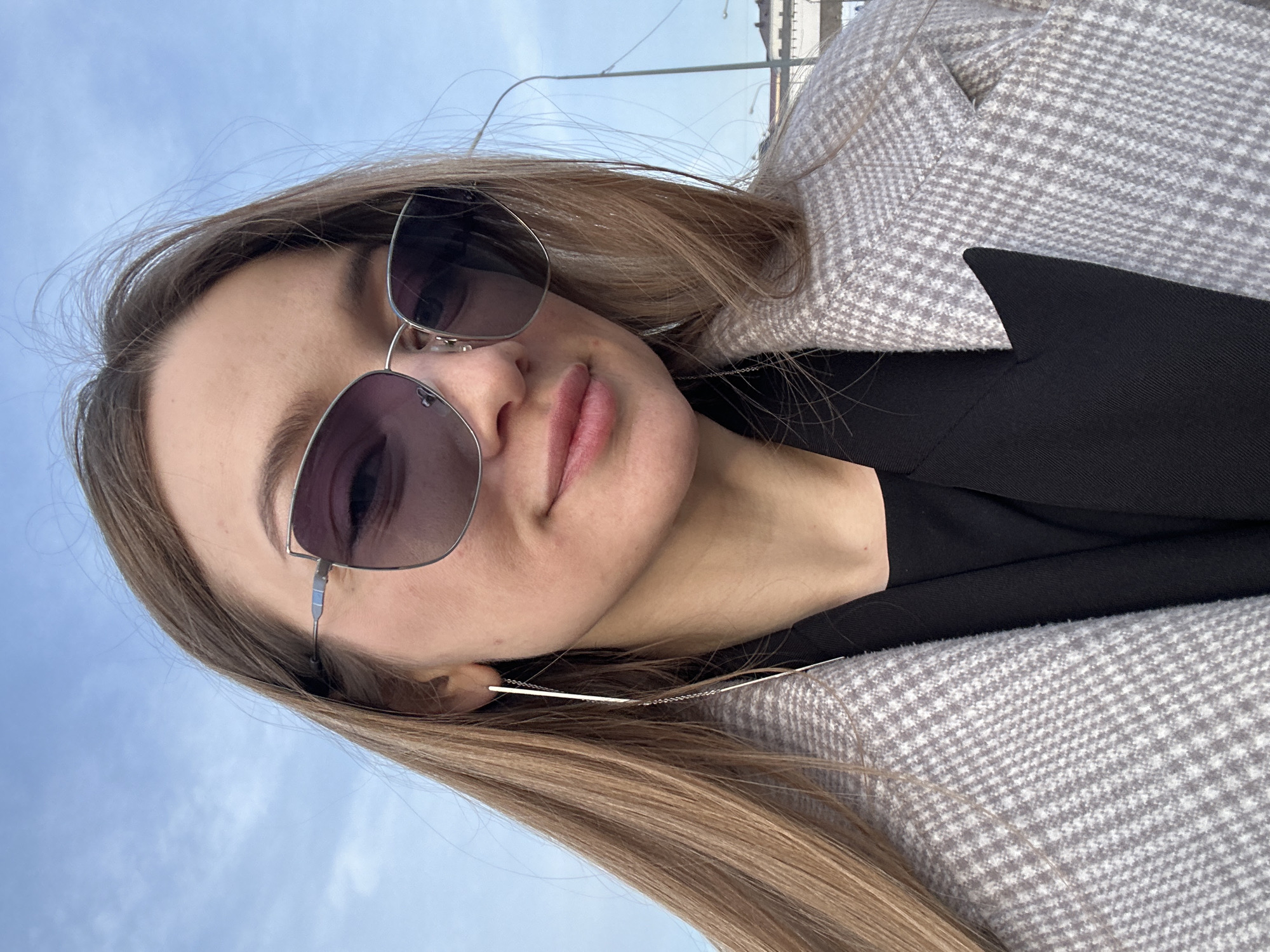 A close-up, head-and-shoulders portrait depicts a female individual outdoors. She has long, light brown hair and is wearing large, grey-tinted sunglasses with thin silver frames featuring a slight cat-eye shape and a horizontal blue accent on the temples. Long, thin, silver-colored threader earrings are visible, extending to her shoulders. Her lips are a natural pink, slightly upturned in a subtle smile. She is dressed in a black top or inner garment, overlaid with a light grey and white houndstooth pattern overcoat. The background consists primarily of a clear to partly cloudy blue sky. To the right, a streetlamp pole with multiple fixtures is partially visible, and in the distance, indistinct urban architectural structures are present. The specific location within the city of Kazan, Russia, is not identifiable from the visual evidence.