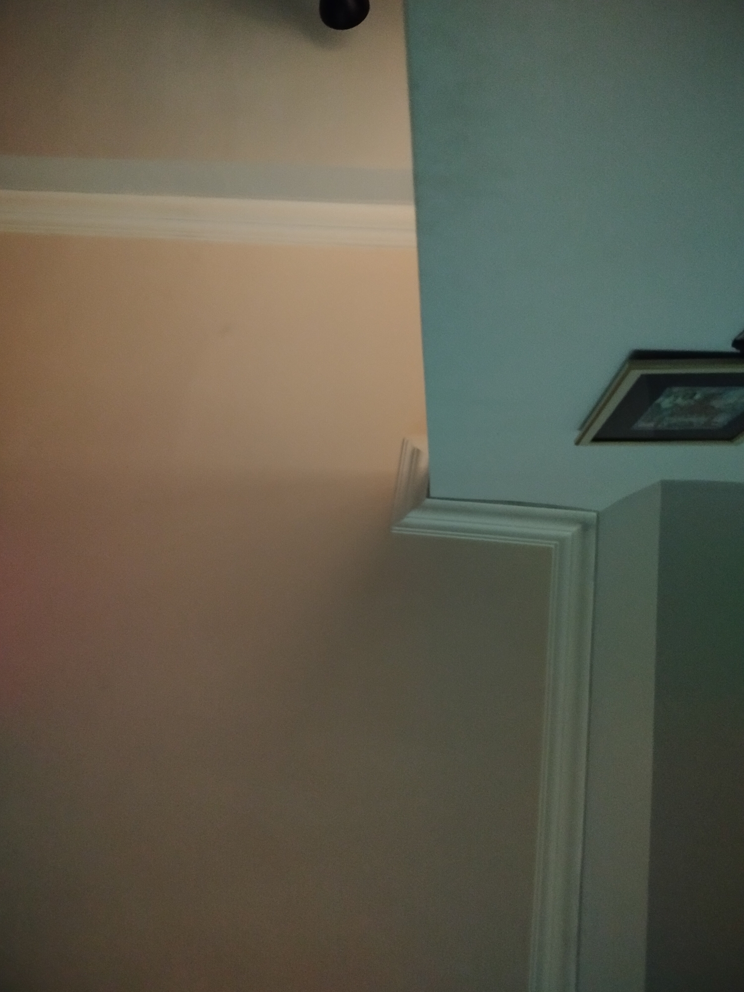 The image shows the intersection of two walls at a corner of a room. The wall to the left is painted in a light peach color with white crown molding at the top. The wall to the right is painted in a light blue color, also with white crown molding where it meets the ceiling. A framed picture hangs on the right wall. The location is not identifiable as Yerevan, Armenia from this image.
