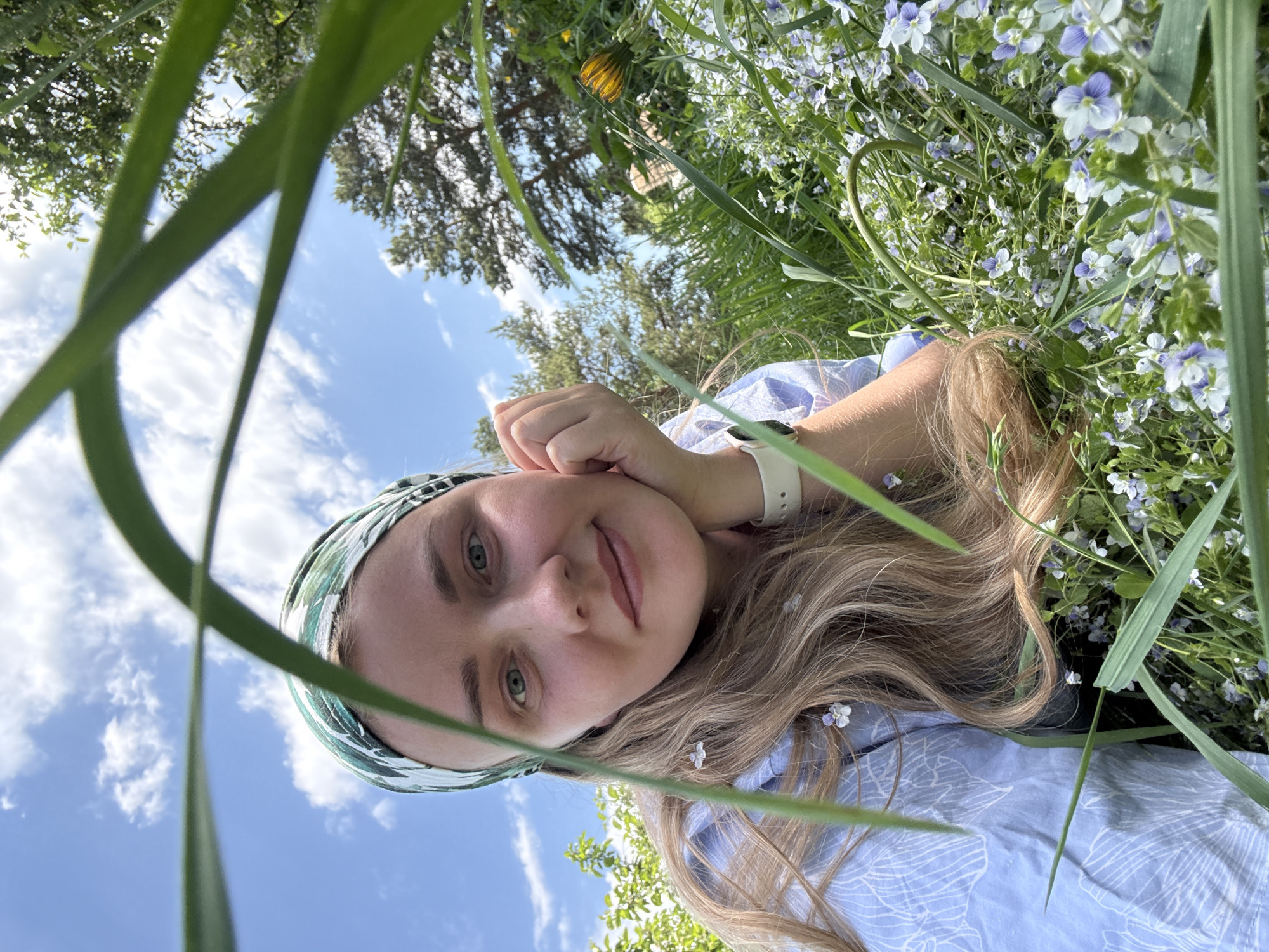A young woman is lying in grass and small blue flowers in what appears to be a park on a sunny day. She wears a patterned headscarf and a light blue shirt. She is propped up on her left hand, wearing a white smartwatch. Grass blades are visible in the foreground, partially obscuring the view. Trees and a blue sky with white clouds are in the background. The location is specified as Chaikovsky, Russia.