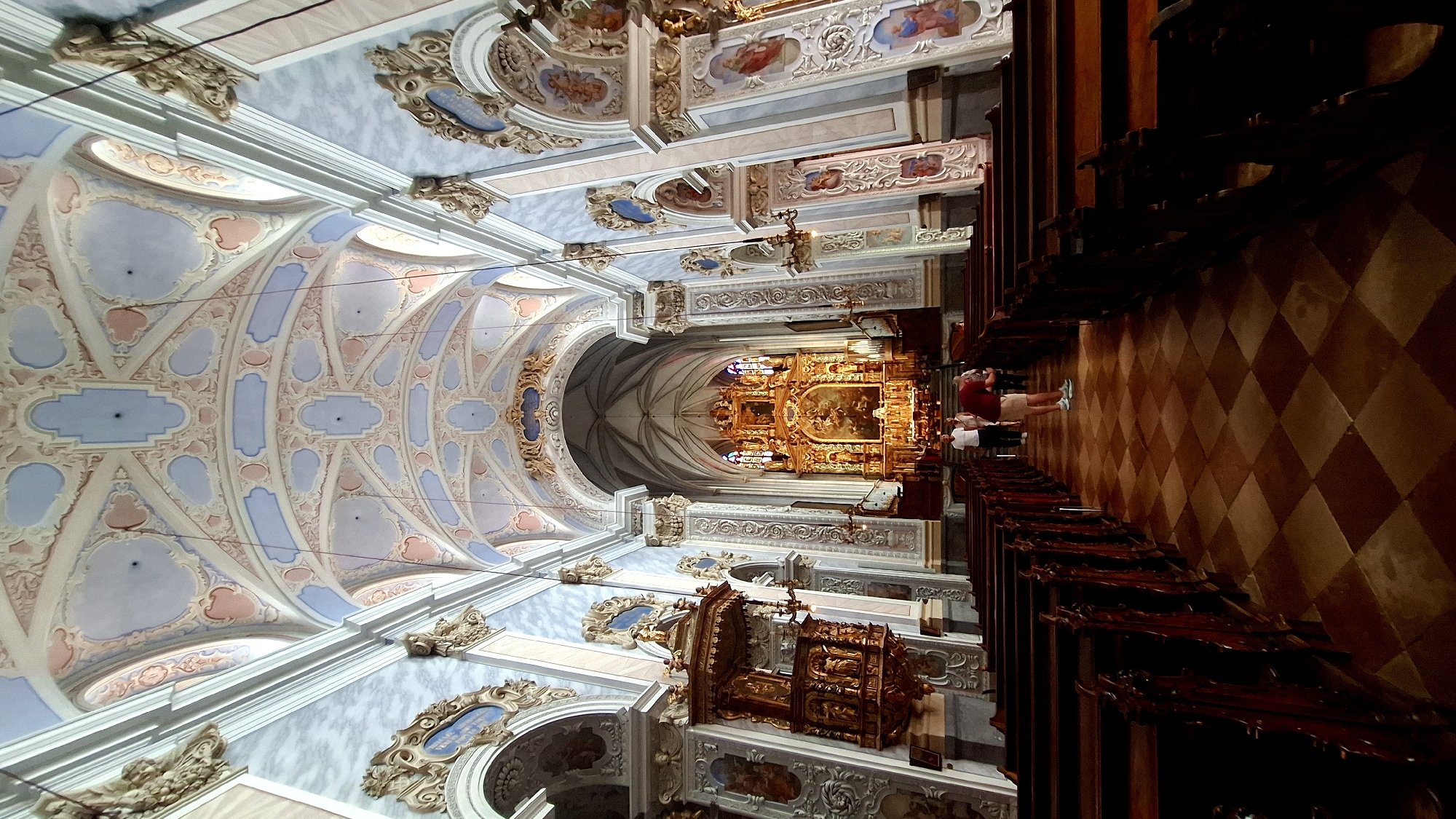 The image shows the interior of the baroque church in Furth bei Göttweig, Austria. The nave is characterized by rows of dark wooden pews on either side, separated by a tiled floor with a diamond pattern in brown and beige. The ceiling is elaborately decorated with stucco work in pastel colors, featuring intricate patterns and oval and rectangular recesses. Light filters in from unseen windows, illuminating the space.
At the far end of the nave is the high altar, a grand gilded structure adorned with paintings and statues, set against a backdrop of stained-glass windows. Flanking the altar are organ pipes. To the left of the nave, a richly carved wooden pulpit is visible, mounted on the wall.
In the middle ground, two individuals are standing near the altar. One is wearing a red shirt and beige shorts, facing away from the camera and appearing to observe the altar. The other, in a white shirt and dark pants, stands beside them. The overall atmosphere is one of solemnity and grandeur, indicative of a place of worship.