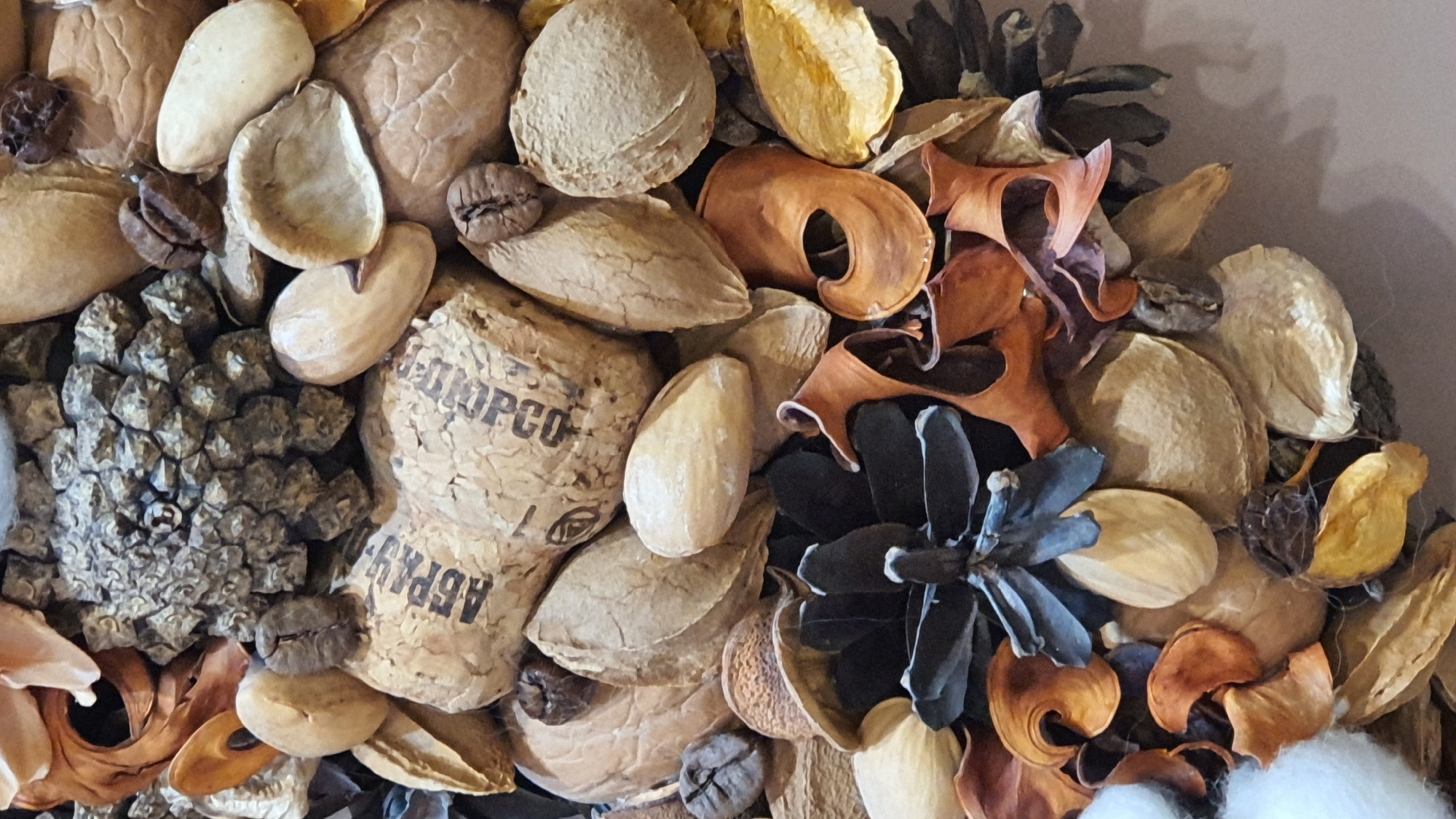The image shows a close-up, overhead view of a potpourri arrangement composed of various natural elements. Prominently featured are dried nuts, seeds, coffee beans, and small pine cones. Some elements appear to be hollowed-out seed pods or fruit shells. There is a piece of what looks like a cork with Cyrillic lettering visible. The arrangement is textural and monochromatic, with shades of brown, beige, and grey dominating. The background is a plain, light-colored surface, likely a wall or table. There are no people or specific landmarks visible that would allow for the identification of a location in Krasnoyarsk, Russia. The scene is static and appears to be an artistic or decorative composition.