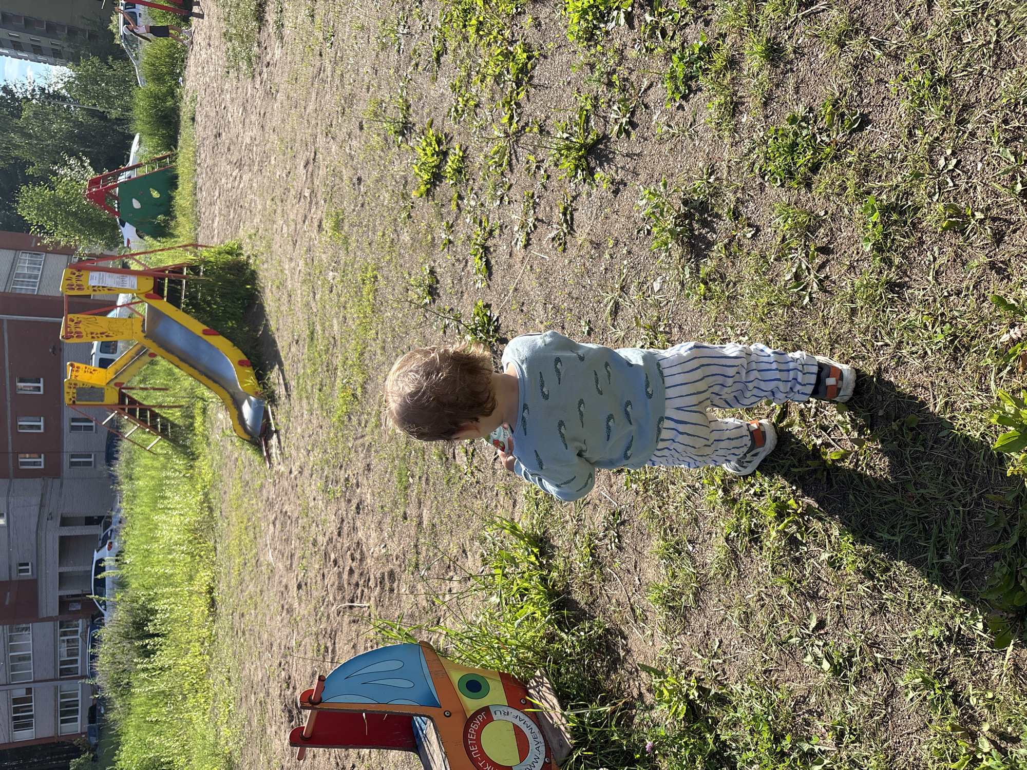 A young child with blond hair stands in a playground, facing away from the camera. The child is wearing a light blue shirt with a pattern of green shapes and striped pants. To the child's left is a brightly colored toy boat. In the background, a yellow and red slide structure and a green climbing wall are visible. The playground is situated on a patch of sandy ground with scattered patches of grass and weeds. In the far background, residential buildings and trees can be seen, suggesting an urban or suburban setting. The lighting indicates it is a sunny day. The image does not contain any information that can definitively identify the location as Chaikovsky, Russia.
