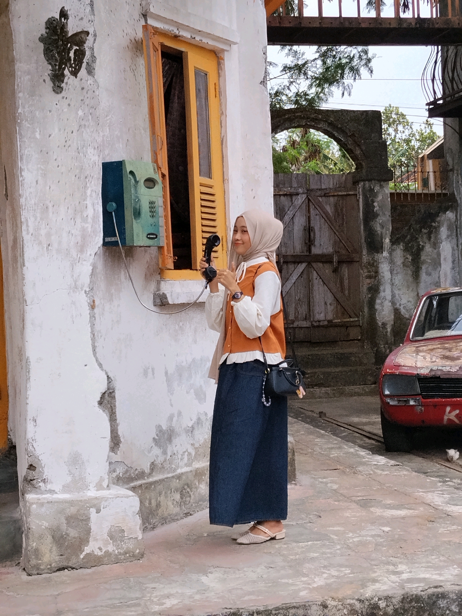 A young woman wearing a hijab and an orange vest over a white top, dark blue skirt, and beige flat shoes, stands in front of a weathered white wall in Sumberrahayu, Indonesia. She is holding a black telephone receiver to her ear, with the cord connected to a green and blue payphone mounted on the wall. The payphone is positioned next to a yellow-framed window with wooden shutters. To the right, an old red car with visible rust and damage is parked. In the background, a wooden gate and a stone archway are visible, along with trees and foliage. The ground is paved with concrete and worn tiles.