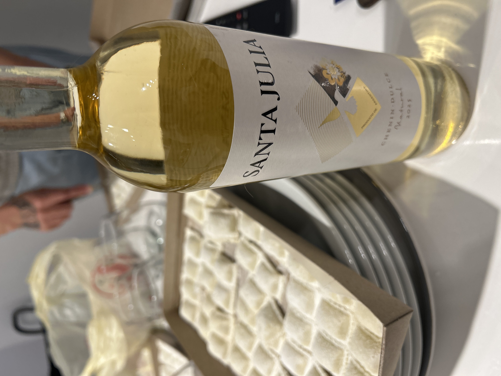 The image shows a bottle of Santa Julia Chenin Dulce Natural 2023 white wine. It appears to be indoors, likely on a table. Near the wine bottle is a box of what appear to be ravioli. Stacked plates are also visible near the ravioli. The label indicates the wine is from Argentina. The scene suggests a meal is being prepared or served. No specific location in Buenos Aires is discernible from the image. A person's arm is partially visible in the background, suggesting someone is nearby.