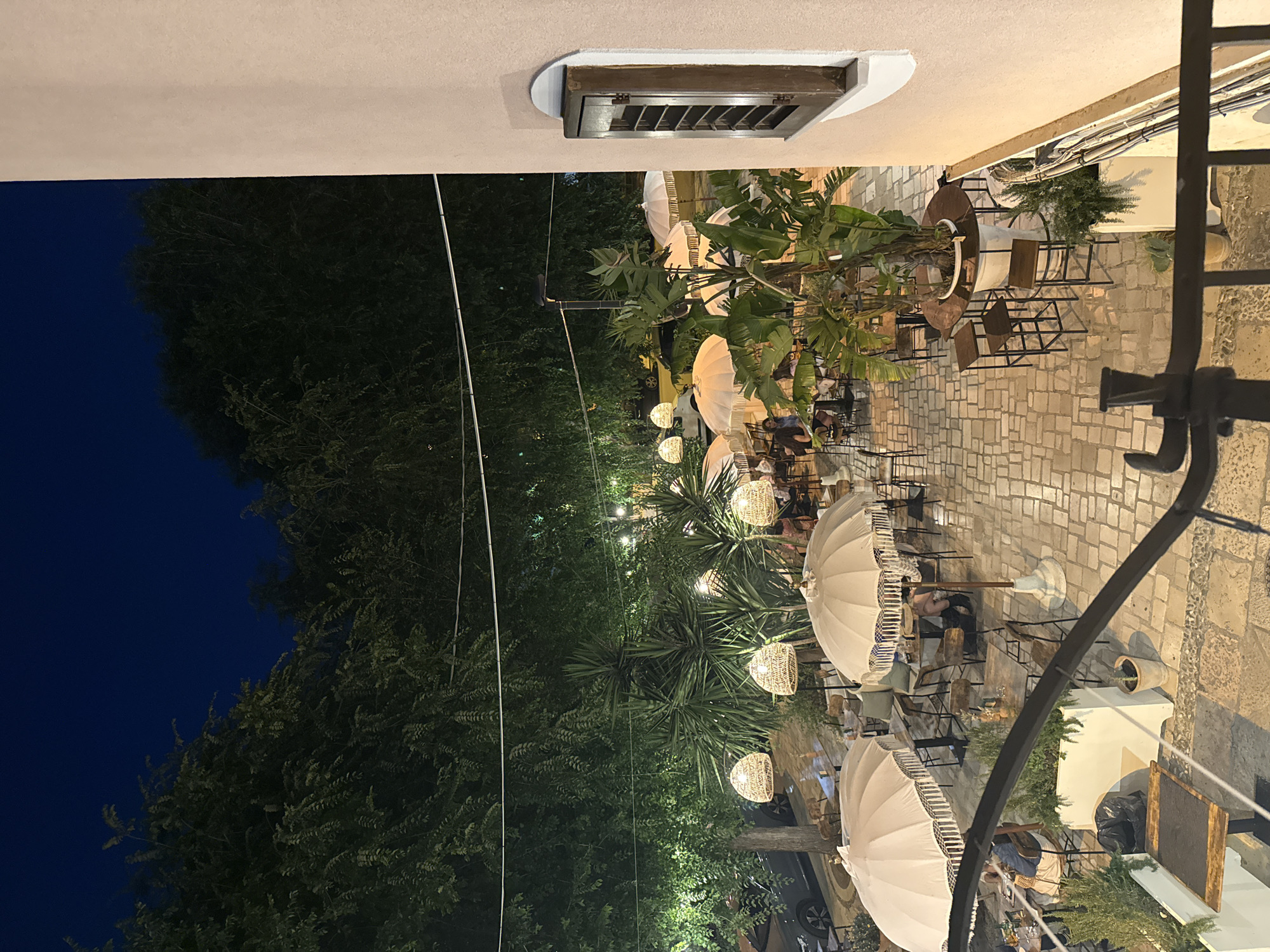 This is an outdoor nighttime scene at a restaurant located in Barletta, Italy. The courtyard of the restaurant is paved with stone tiles and features several tables and chairs for outdoor dining. Large, white, fringed umbrellas provide shade. Woven pendant lights hang from trees above, and string lights are also visible, casting a warm glow over the area. Lush greenery, including large palm-like plants and trees, surrounds the courtyard. In the background, a car is parked on a street. A building with a light-colored facade and a window with wooden shutters is visible on the right side of the frame. A few patrons are seated at tables, engaged in dining or conversation. The overall atmosphere is that of a relaxed, open-air dining establishment.